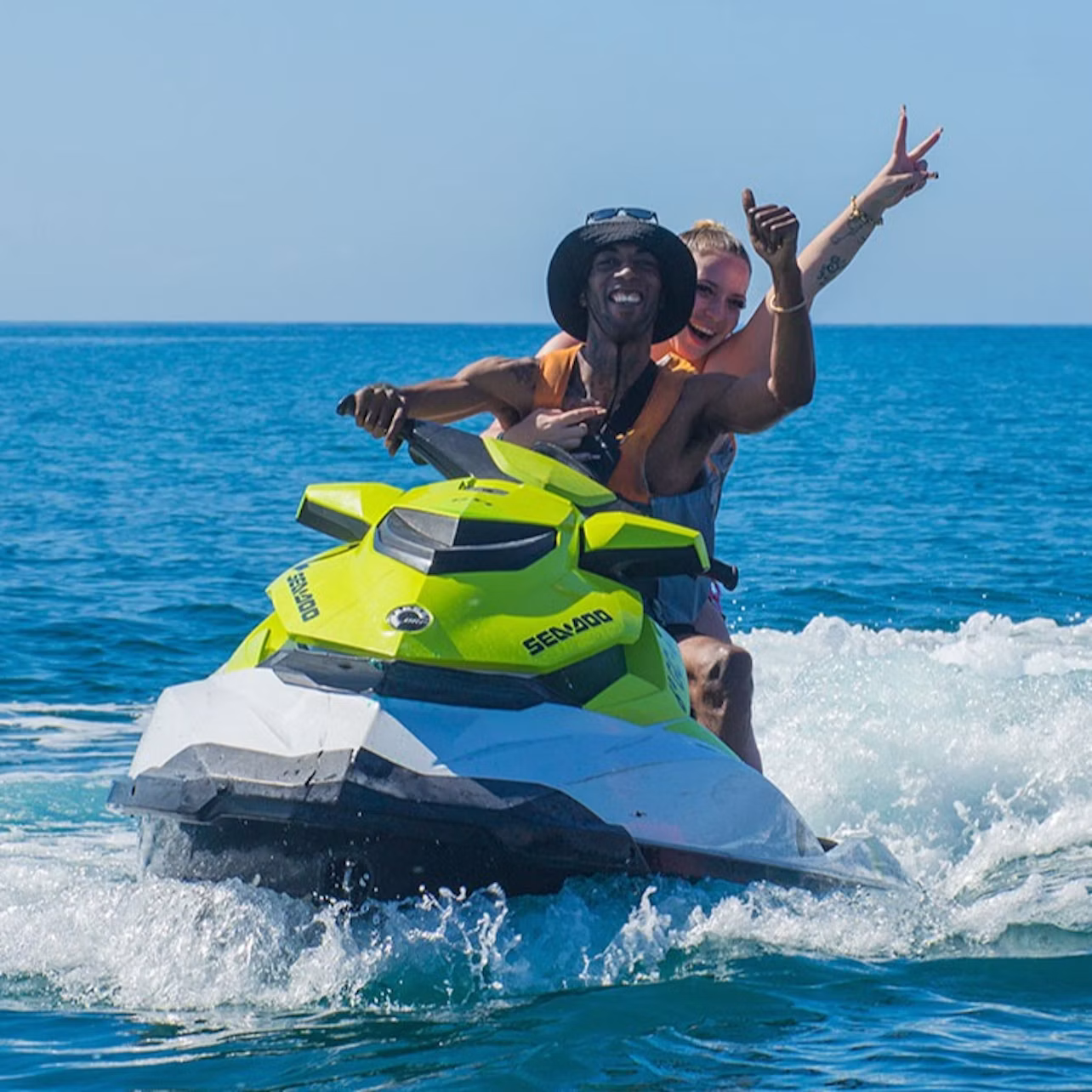 Two people on a yellow and white jet ski riding on blue water, with the passenger making a peace sign and the driver smiling.