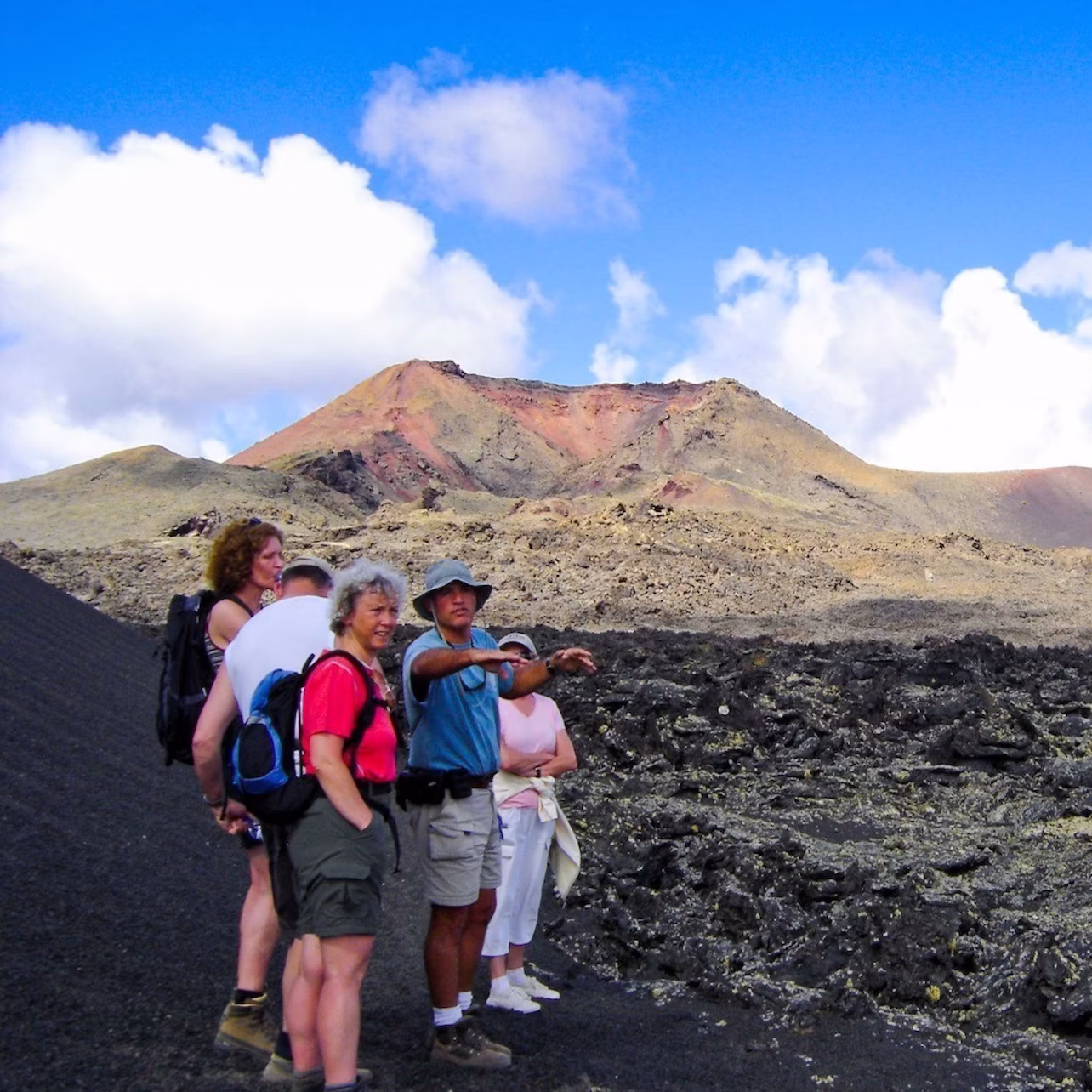 A group of people wearing outdoor attire stand on rocky terrain with a mountain and clear blue sky in the background.