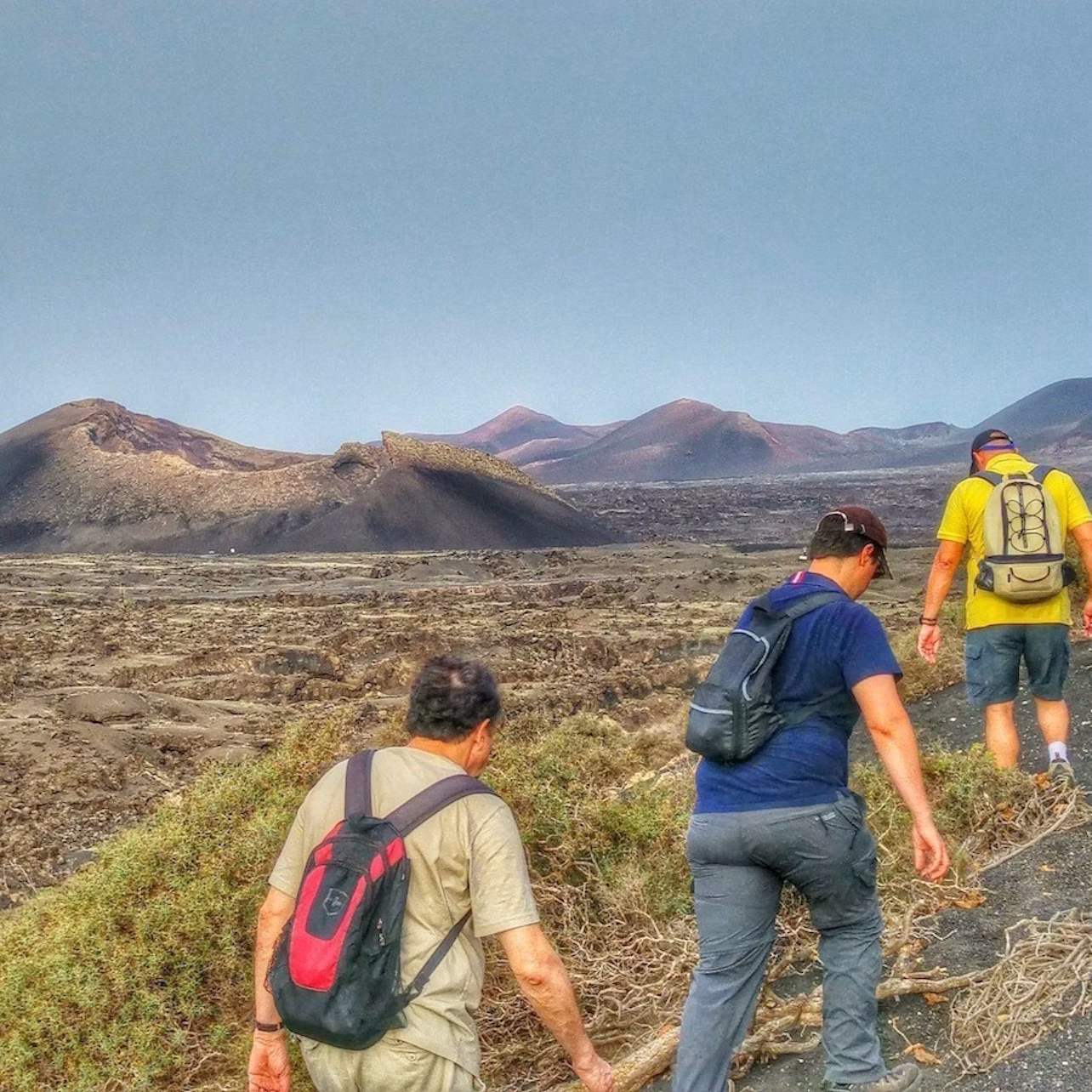 Three hikers with backpacks walking on a rocky terrain with volcanic hills and a clear sky in the background.