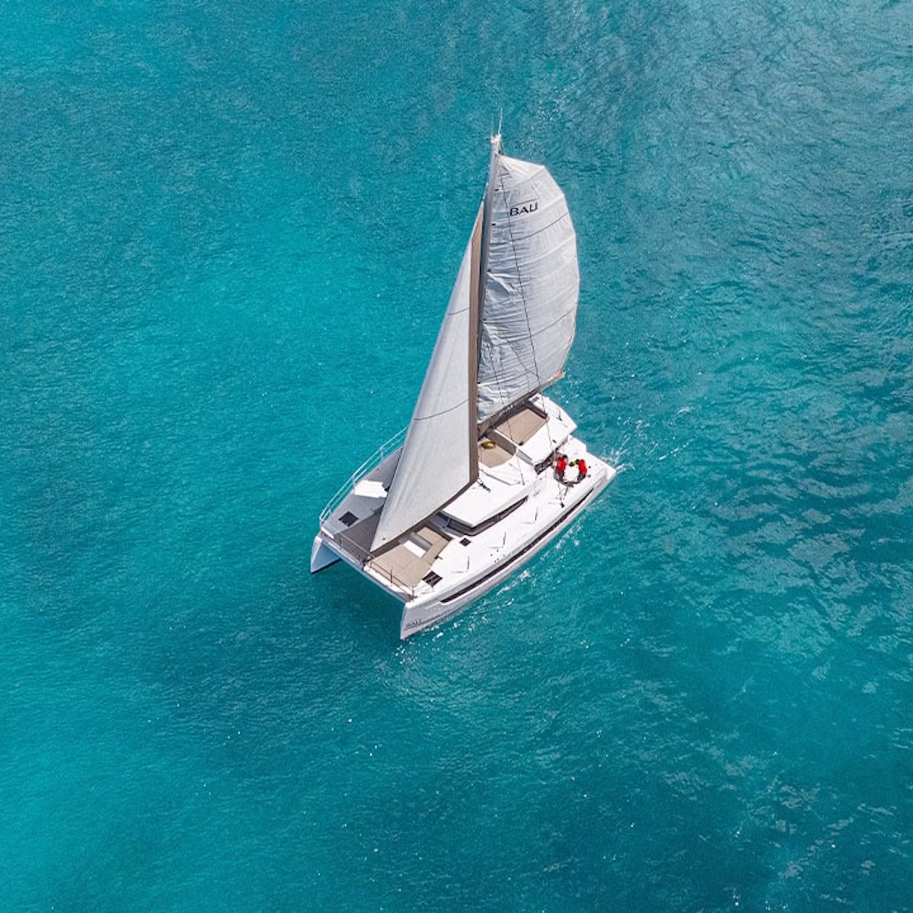 A white sailboat with one sail glides on clear, turquoise water, viewed from above.