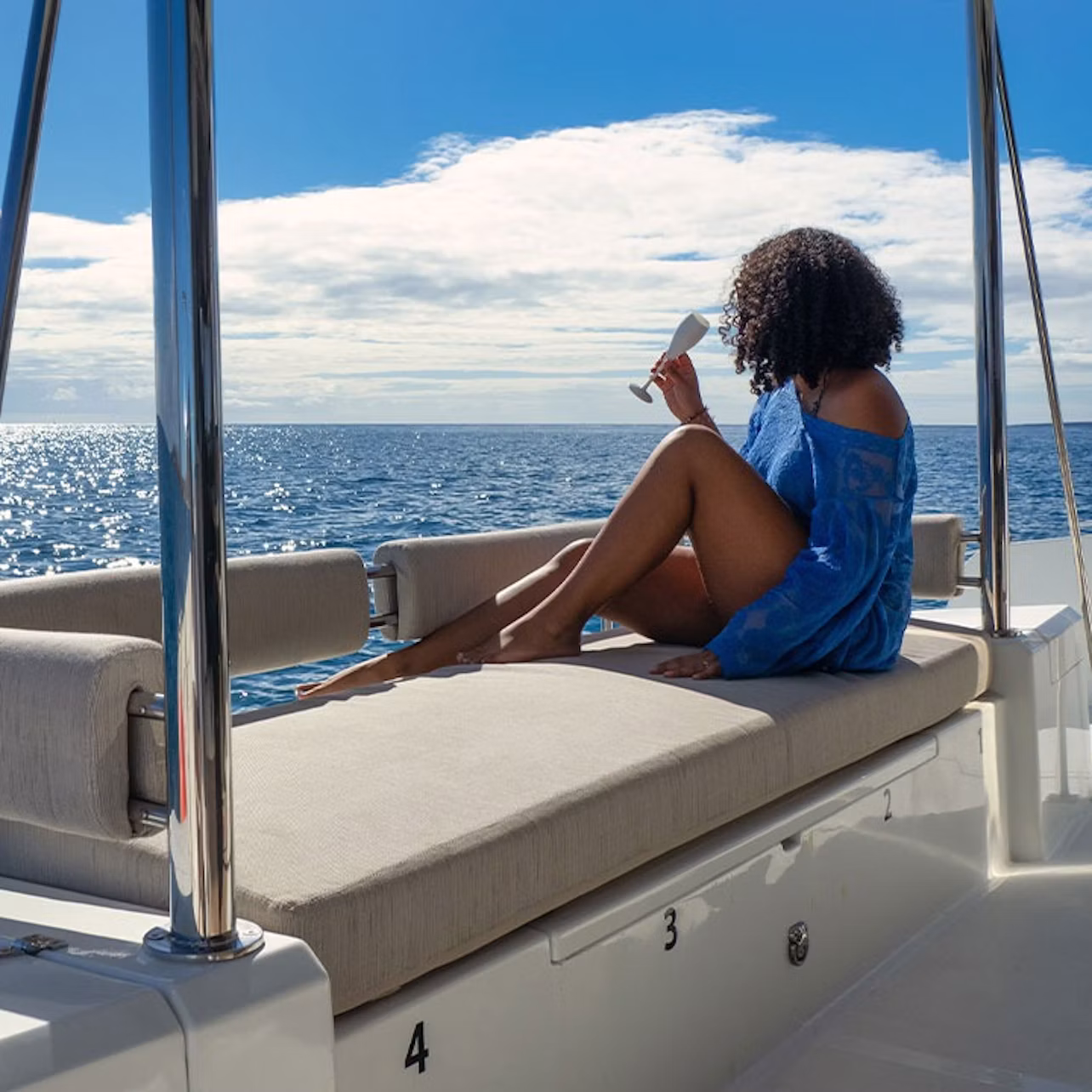 A person with curly hair in a blue cover-up lounges on a boat's cushioned seat, holding a drink and gazing at the ocean under a partly cloudy sky.