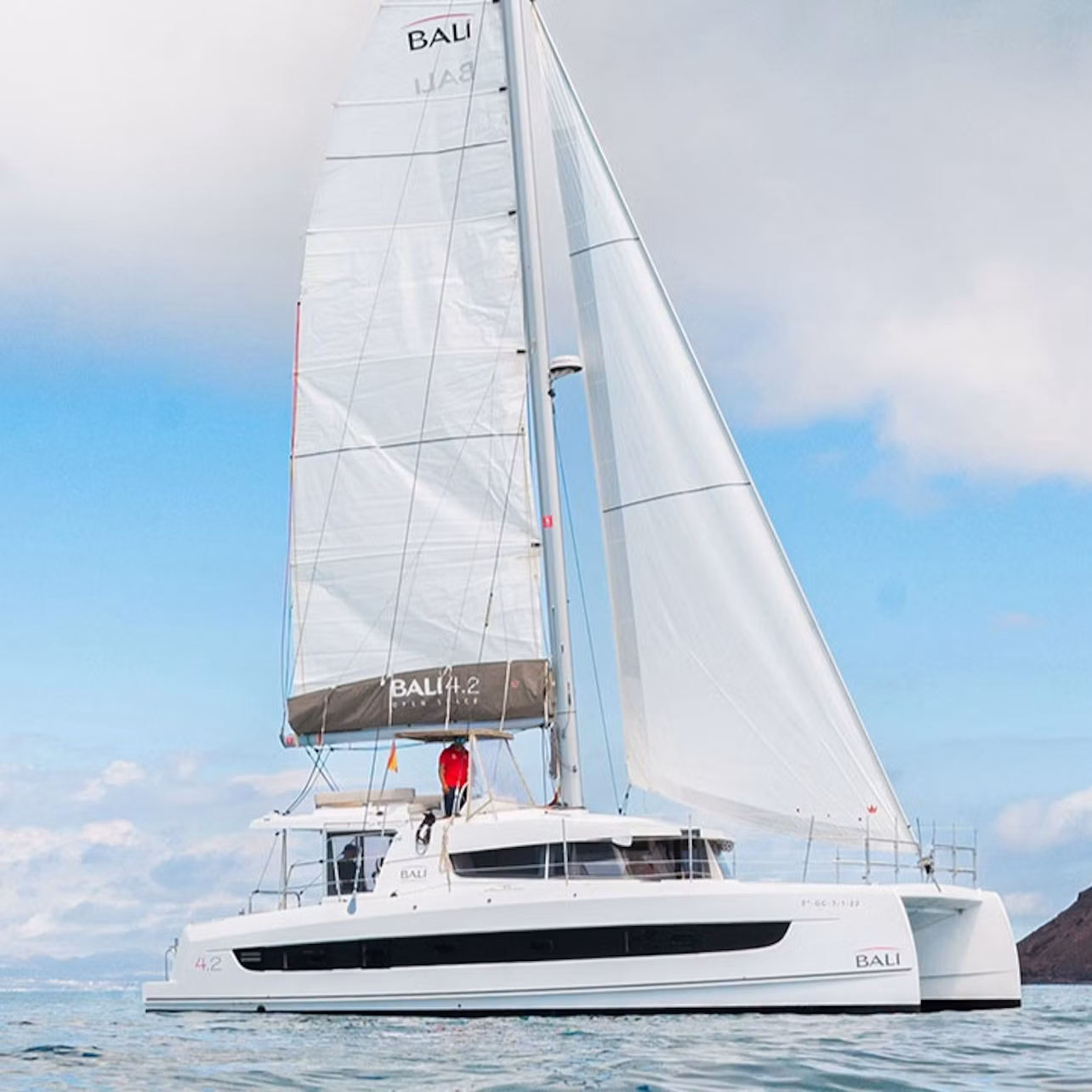A white catamaran with large sails and a person in a red jacket on the deck, sailing on calm water under a cloudy sky.