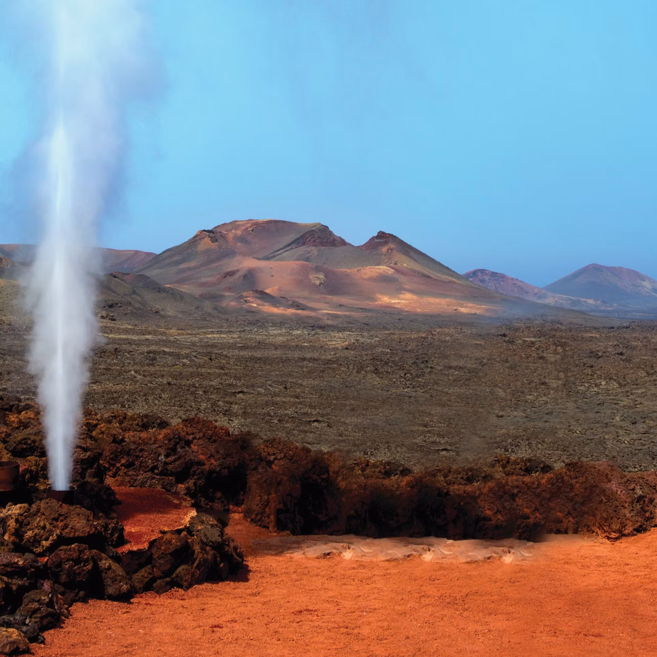 Steam geyser erupts from rocky terrain with red soil, set against a backdrop of barren, mountainous landscape under a clear blue sky.