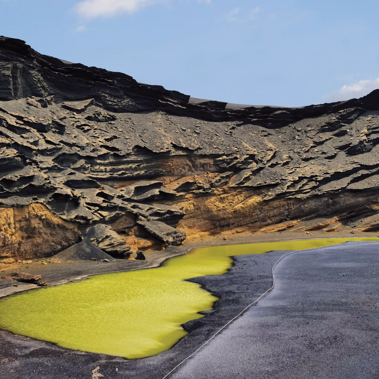 Jagged black and brown rocks surround a small, bright green sulfuric lake under a clear blue sky.