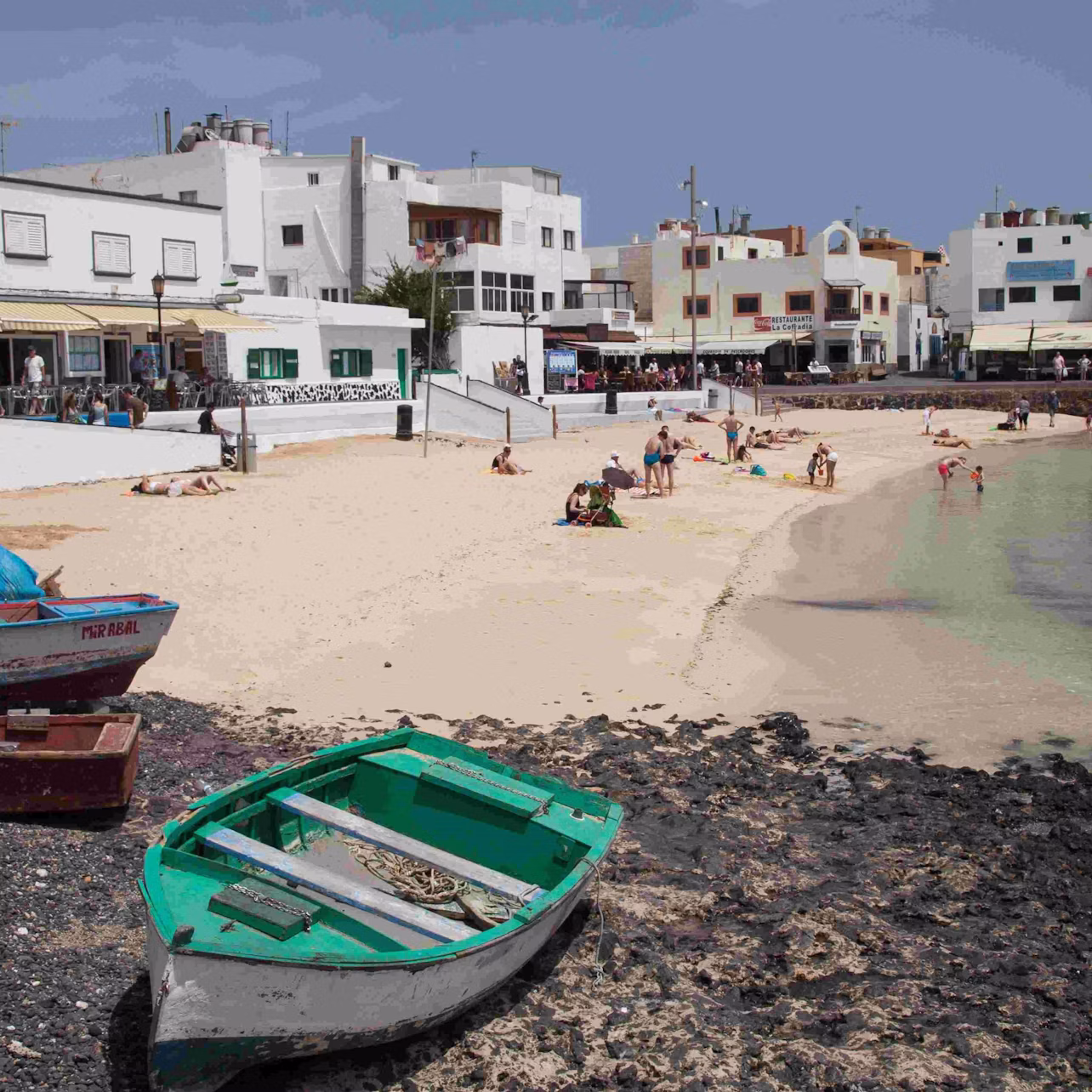A small beach with people sunbathing and swimming, flanked by white buildings. Two rowboats on rocky shore in the foreground.