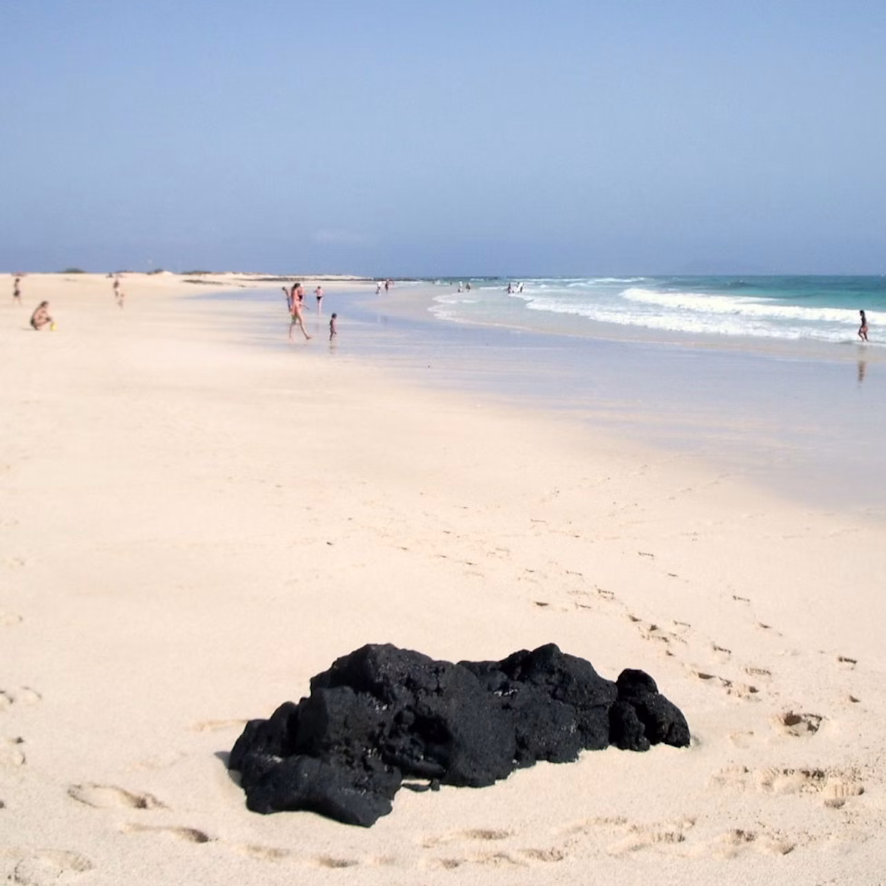 People scattered along a sandy beach with gentle waves; a pile of black rocks in the foreground.