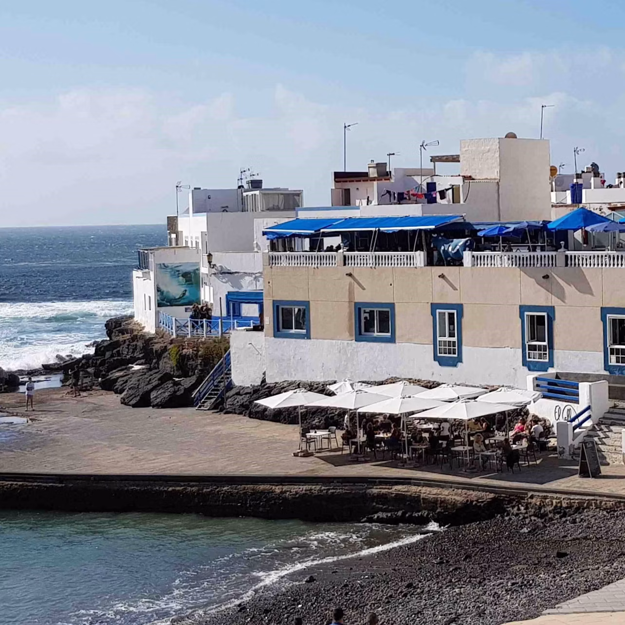 Seaside building with blue trim, outdoor café with white umbrellas on patio, rocky coastline, and waves in the background.