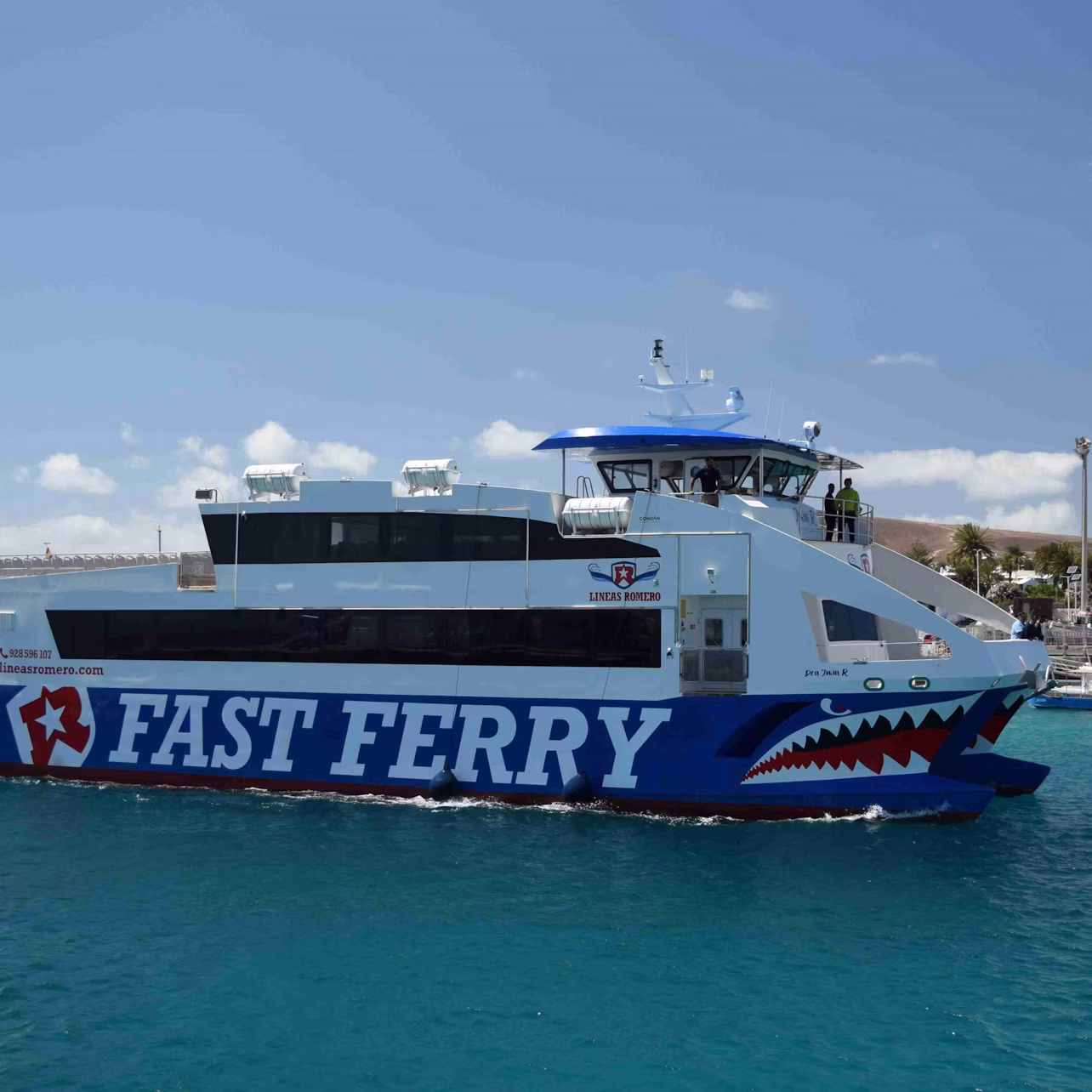 A blue and white ferry labeled "FAST FERRY" with shark graphics sails on clear blue water under a partly cloudy sky.