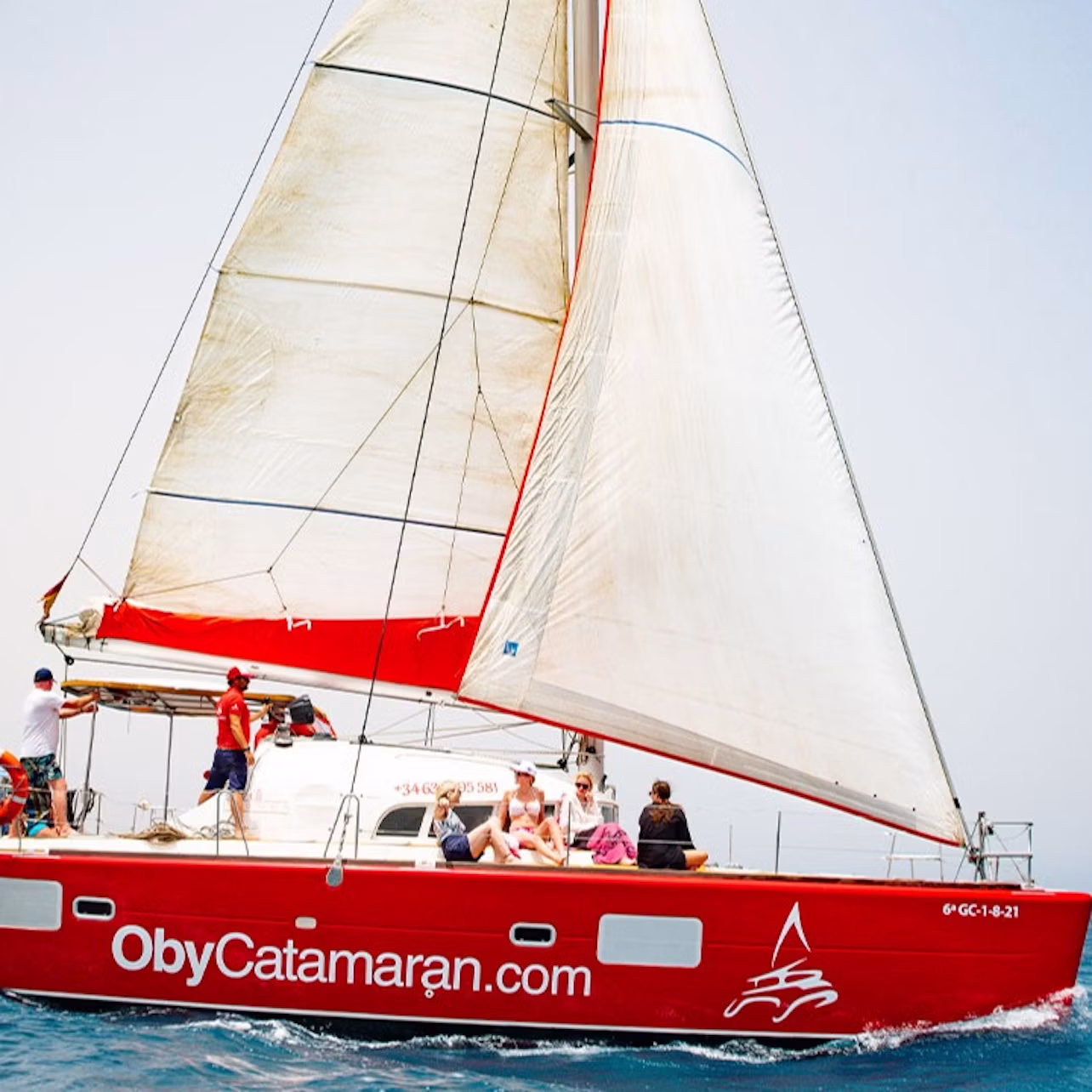 A red catamaran named "ObyCatamaran.com" sails on the sea with people on deck under white sails.