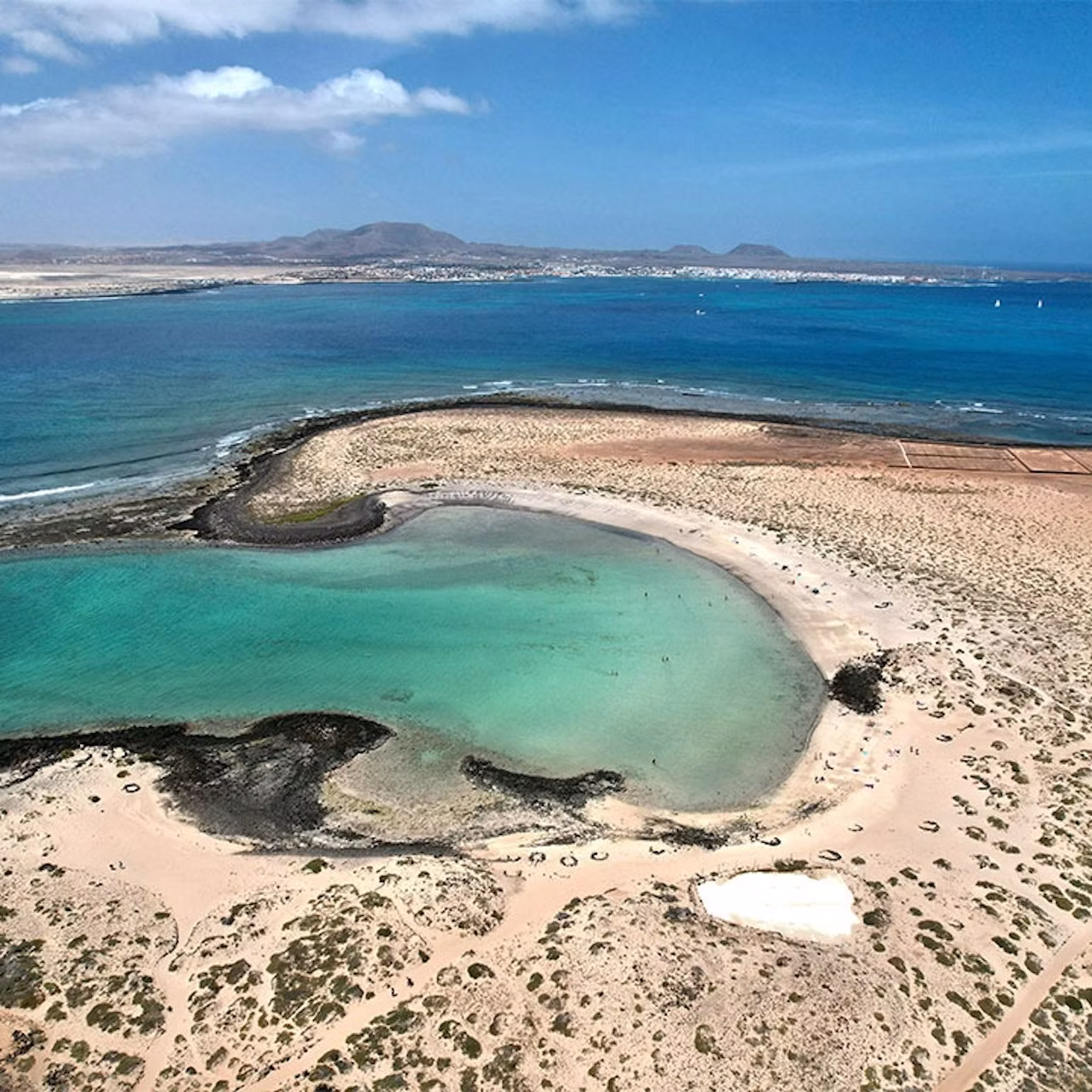 Aerial view of a coastal landscape with turquoise lagoons, sandy beaches, rocky areas, and distant mountains under a blue sky.