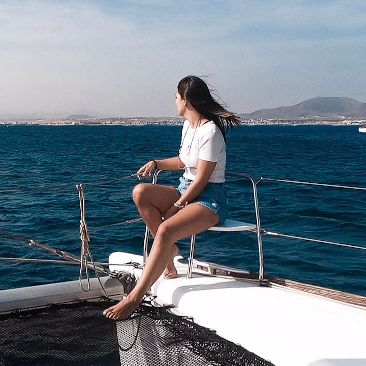 Person sitting on a sailboat, looking out at the blue ocean with mountains in the background.