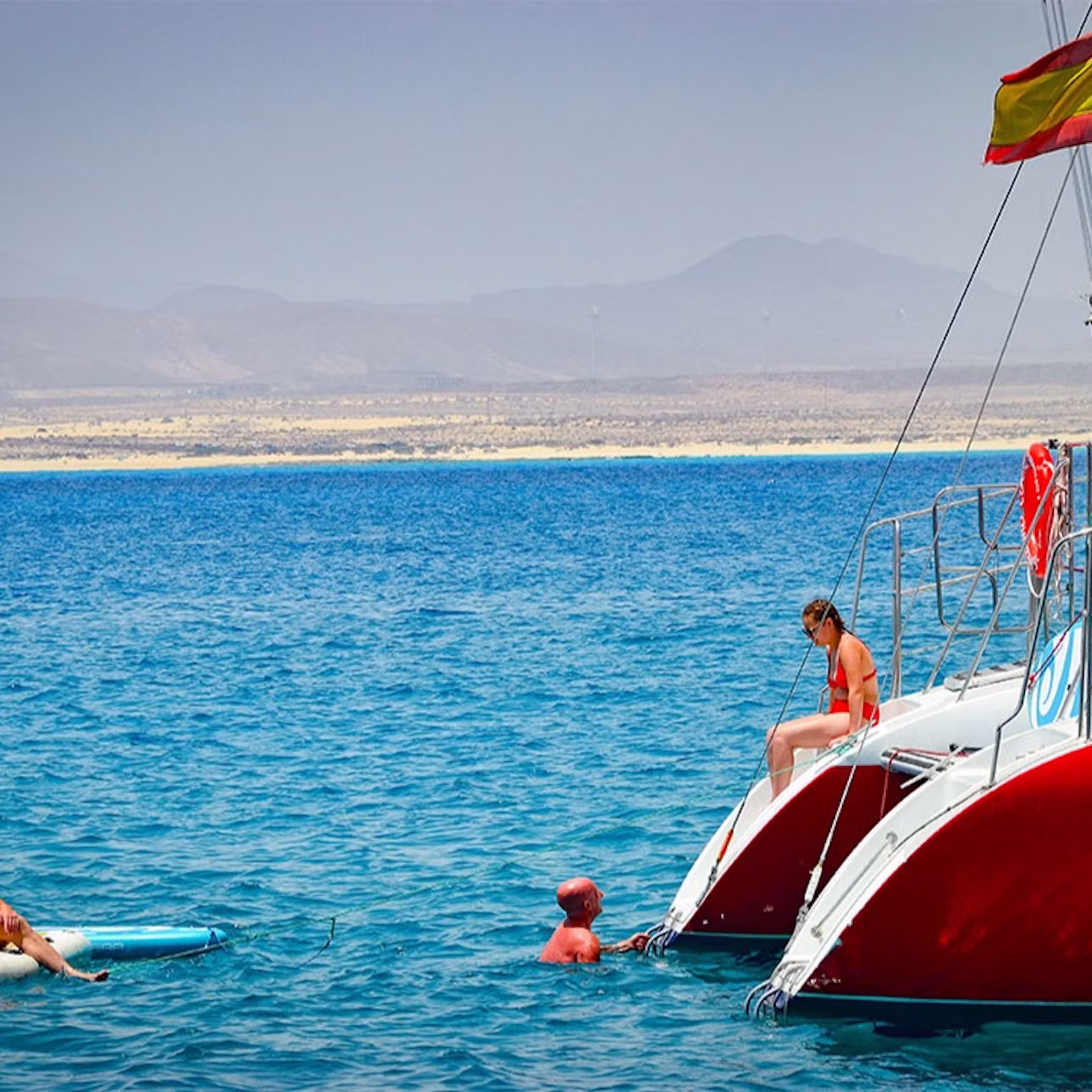 People enjoying the water near a boat with red hulls and a flag, against a backdrop of mountains and clear blue sea.