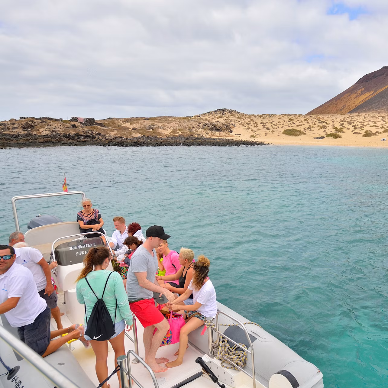 A group of people on a boat near a rocky and sandy coastline with clear blue water under a partly cloudy sky.
