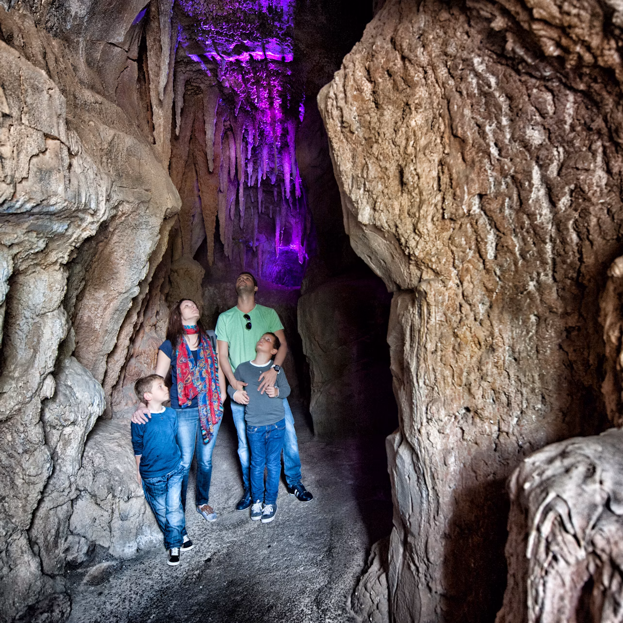 A family of four explores a rocky cave illuminated by purple light, looking up at the stalactites above.