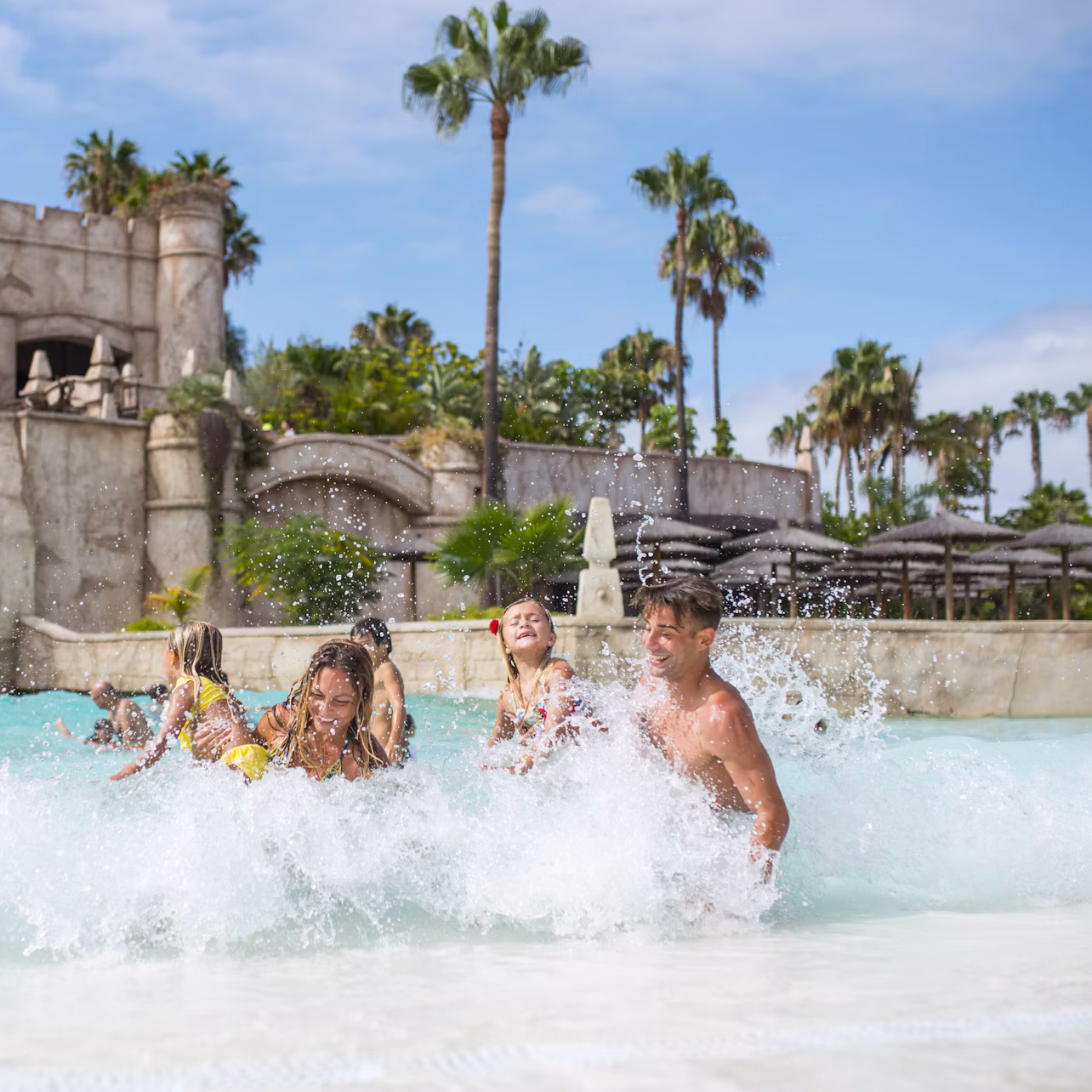 A family enjoying splashing in a shallow pool with a castle-like structure and palm trees in the background.