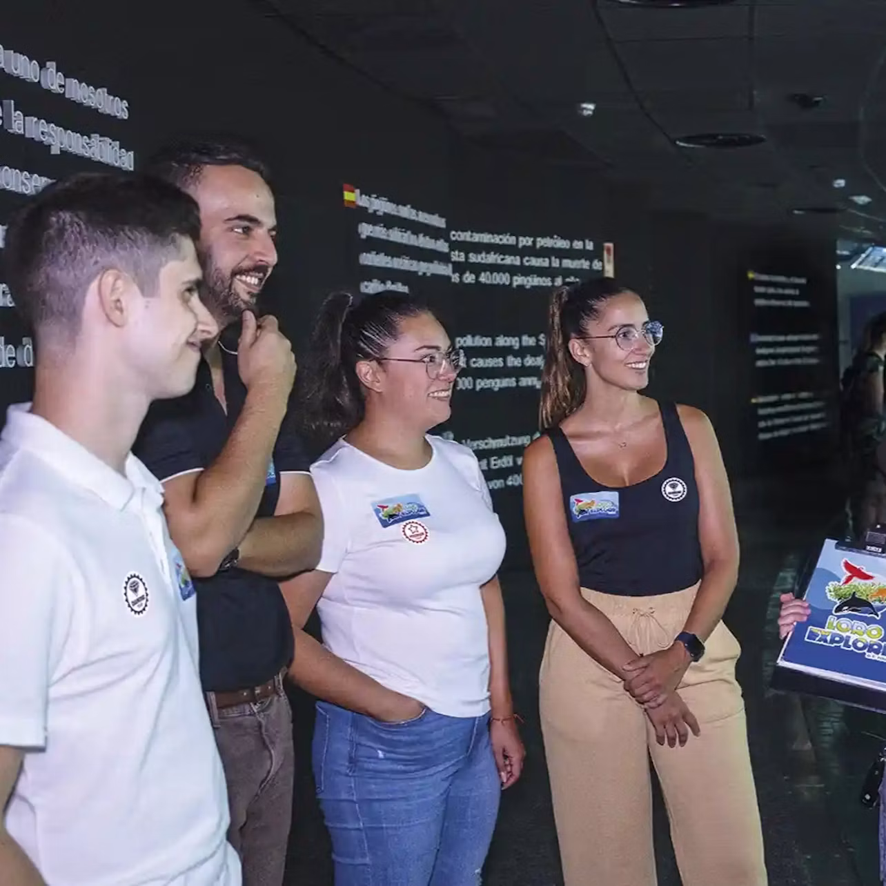 Four people stand indoors, smiling and talking near a display with text. They wear casual clothing with badges.