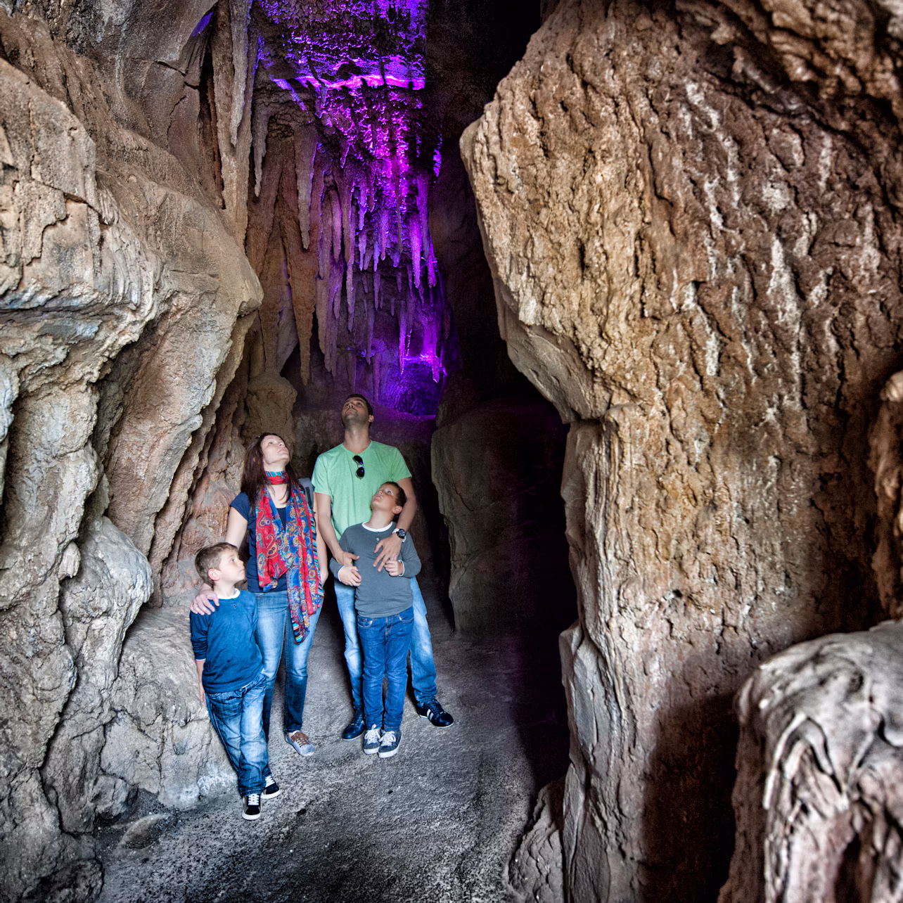 A family of four explores a rocky cave illuminated by purple light, looking up at the stalactites above.