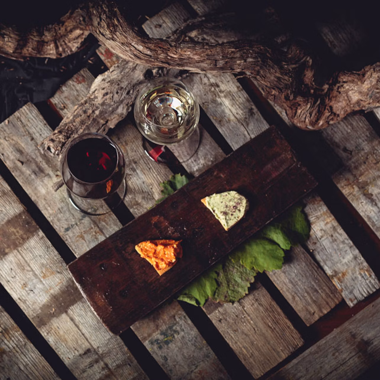 Two glasses of wine and two cheese pieces on a wooden board, set on a rustic wooden surface with a branch overhead.