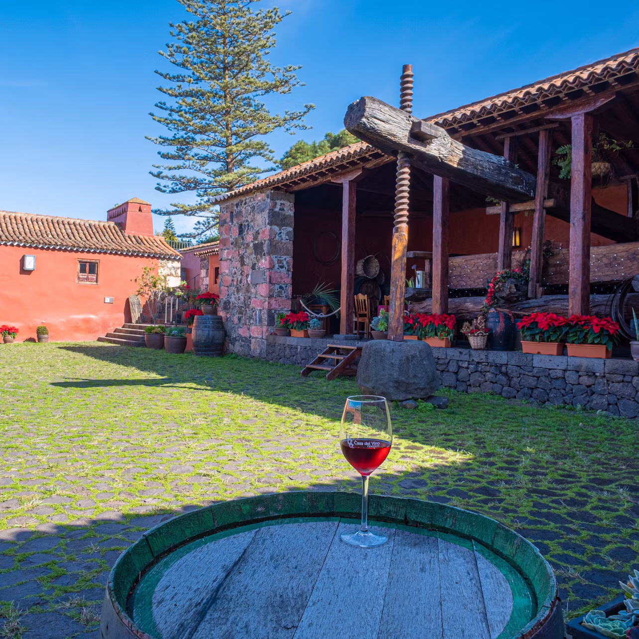 Glass of red wine on a wooden barrel in a courtyard with stone buildings, potted plants, and a traditional wine press.