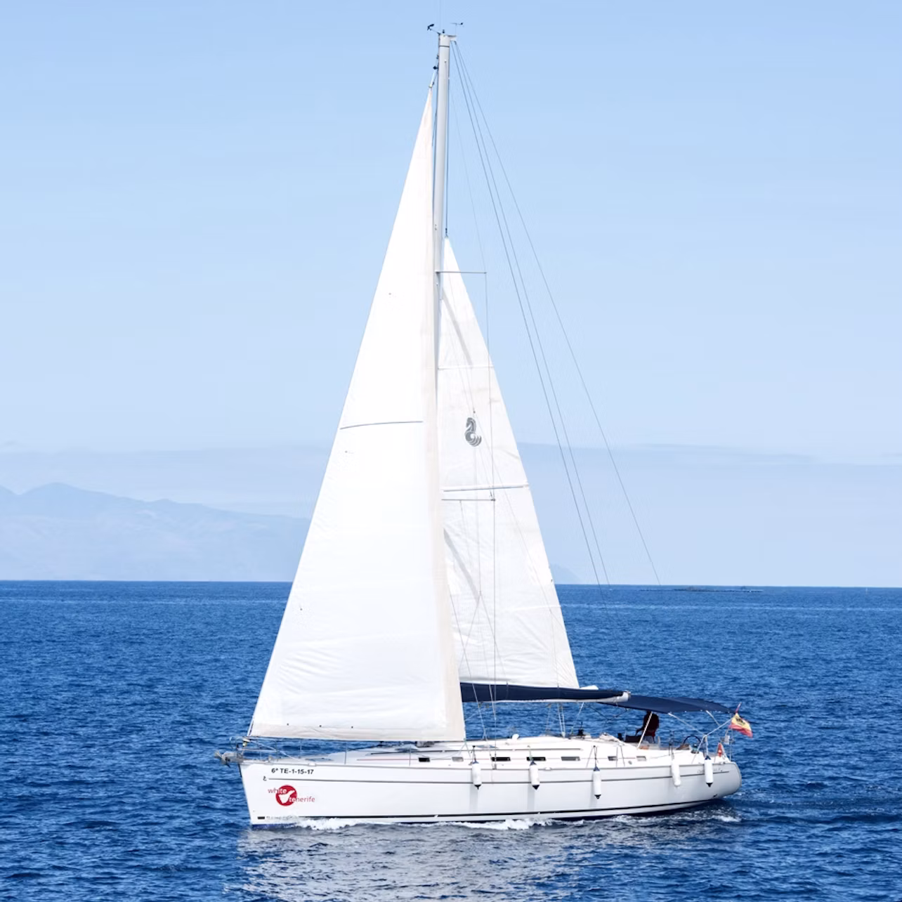 A white sailboat with large sails glides on a calm, blue ocean under a clear sky with distant mountains on the horizon.