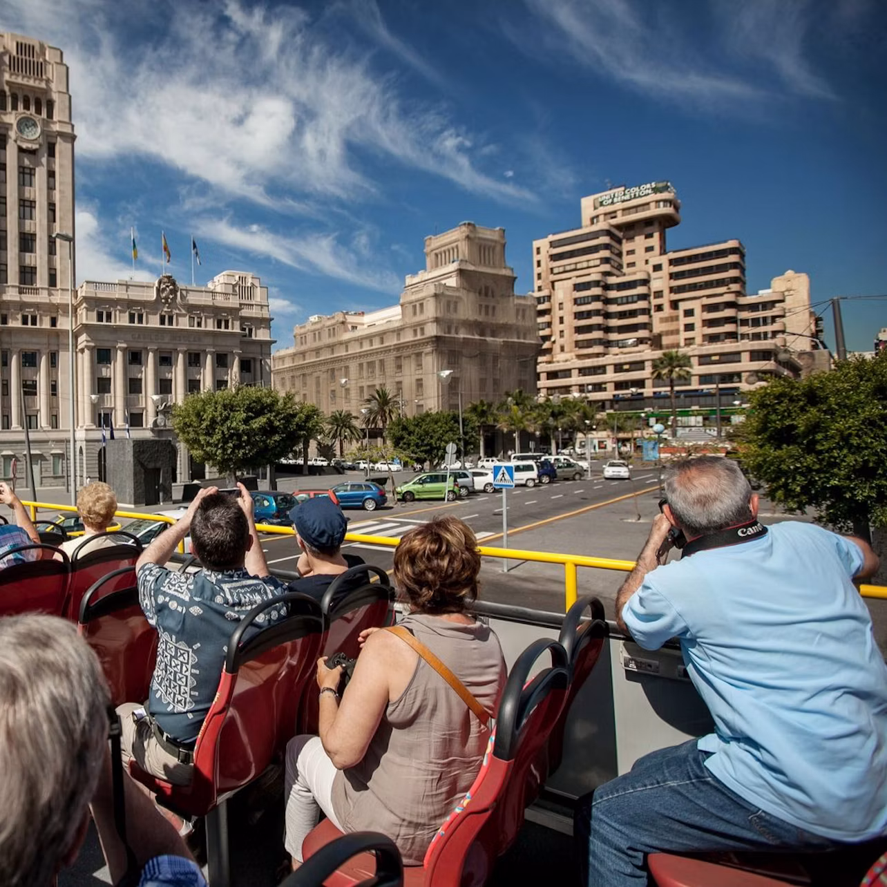 People seated on an open-top tour bus, taking photos of large historic buildings under a partly cloudy blue sky.