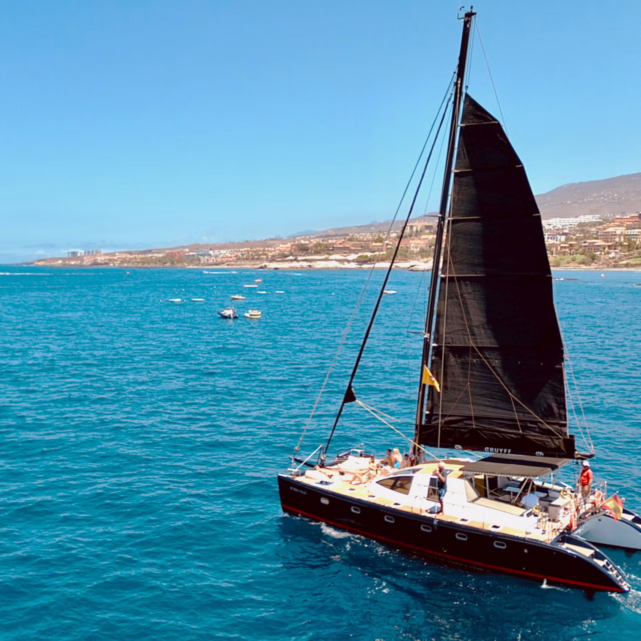 A sailboat with a black sail on a clear blue sea, with a coastline visible in the background under a sunny sky.