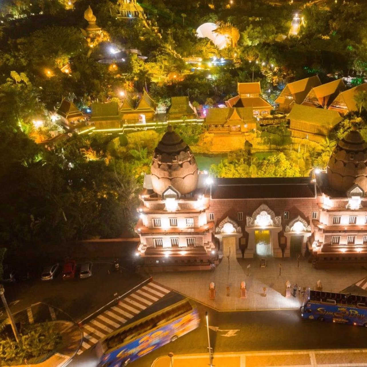 Aerial view of a brightly lit, ornate building at night with lush greenery and various illuminated structures in the background.