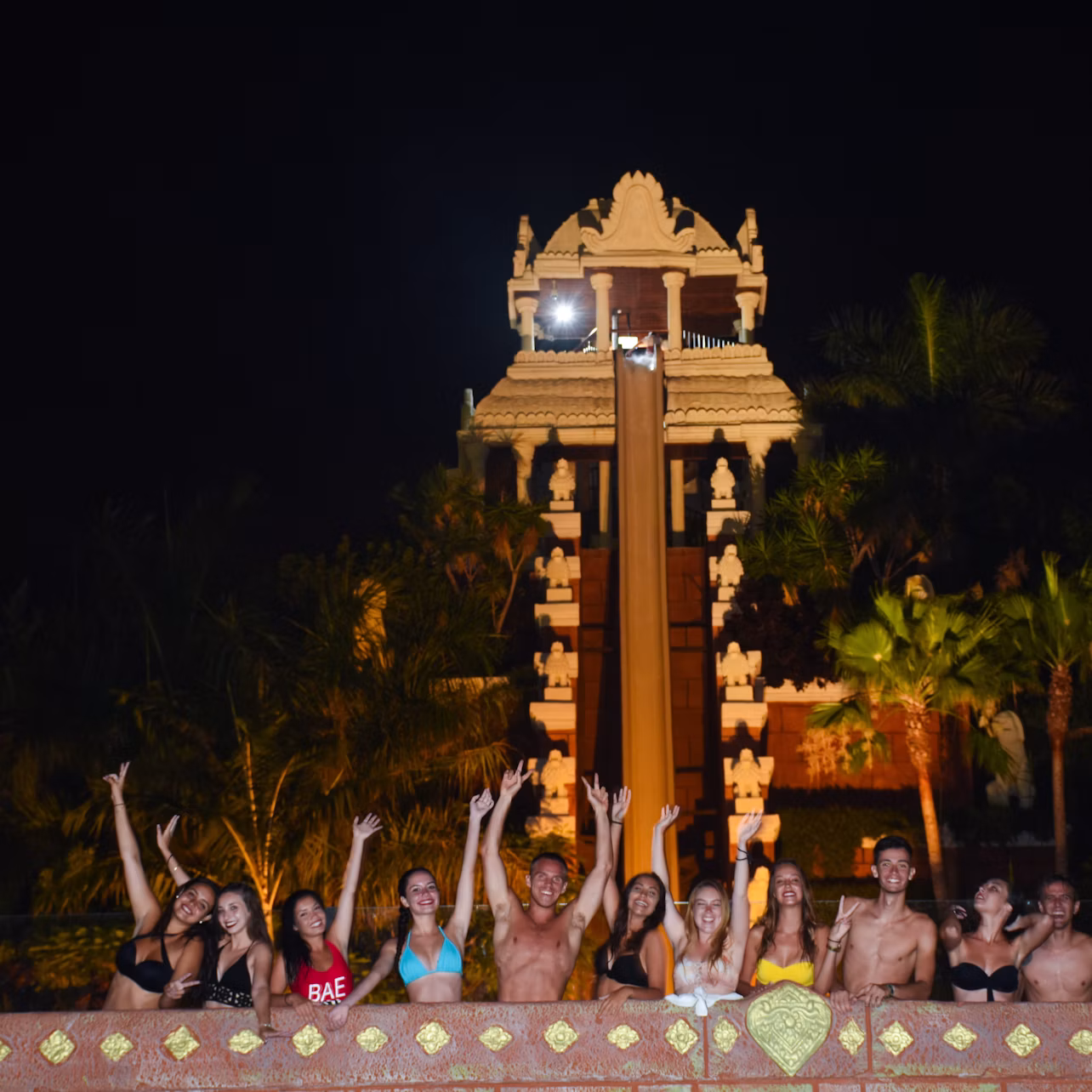 A group of people in swimwear stand in front of a tall, ornate water slide at night, raising their arms and smiling.