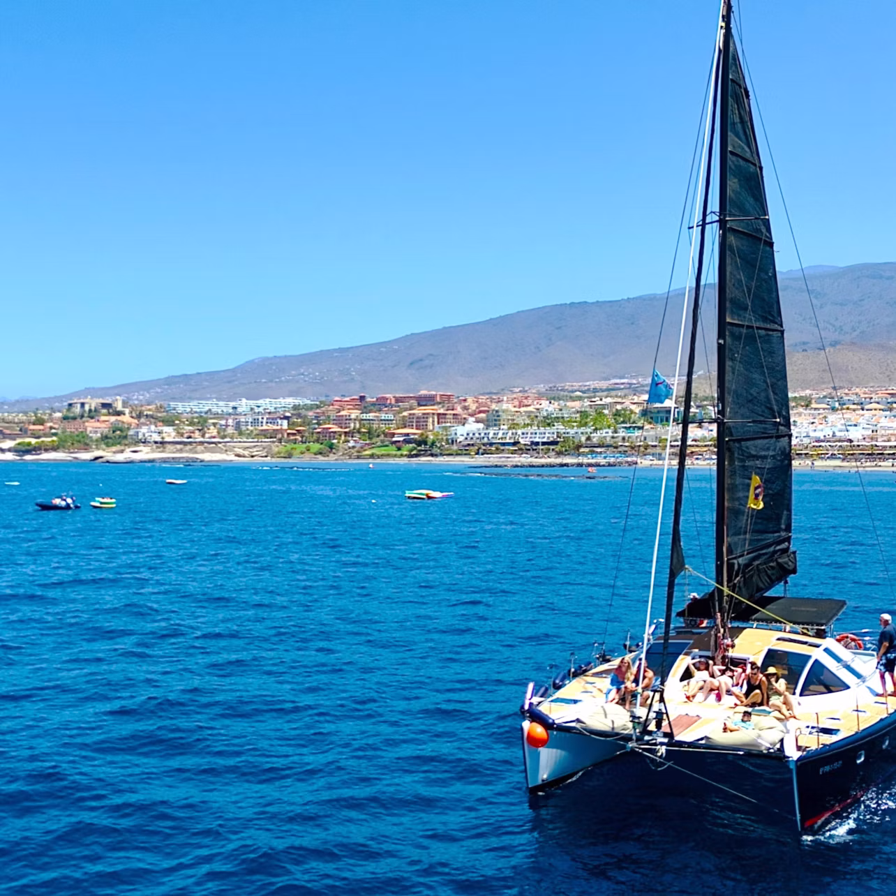 A sailboat with passengers on a calm blue ocean, with a coastal town and hills visible in the background under a clear sky.