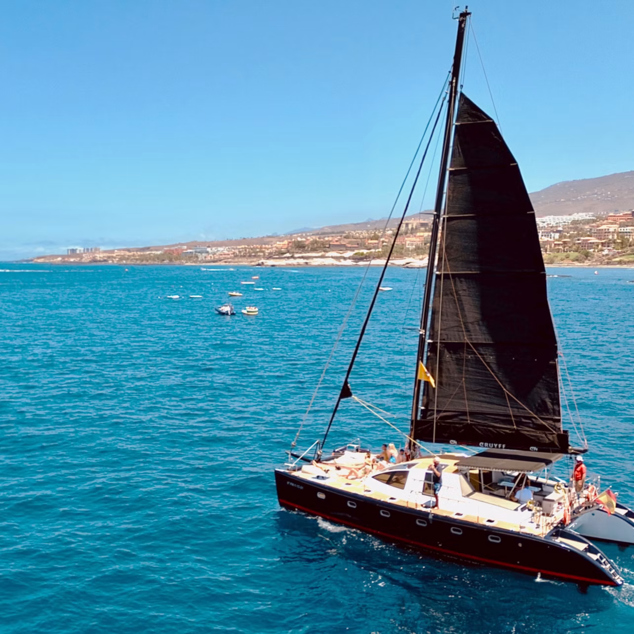 A sailboat with a black sail on a clear blue sea, with a coastline visible in the background under a sunny sky.