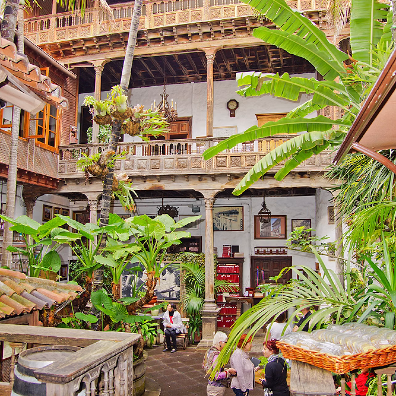 Lush courtyard with people, surrounded by plants and multi-level wooden balconies featuring detailed architecture.