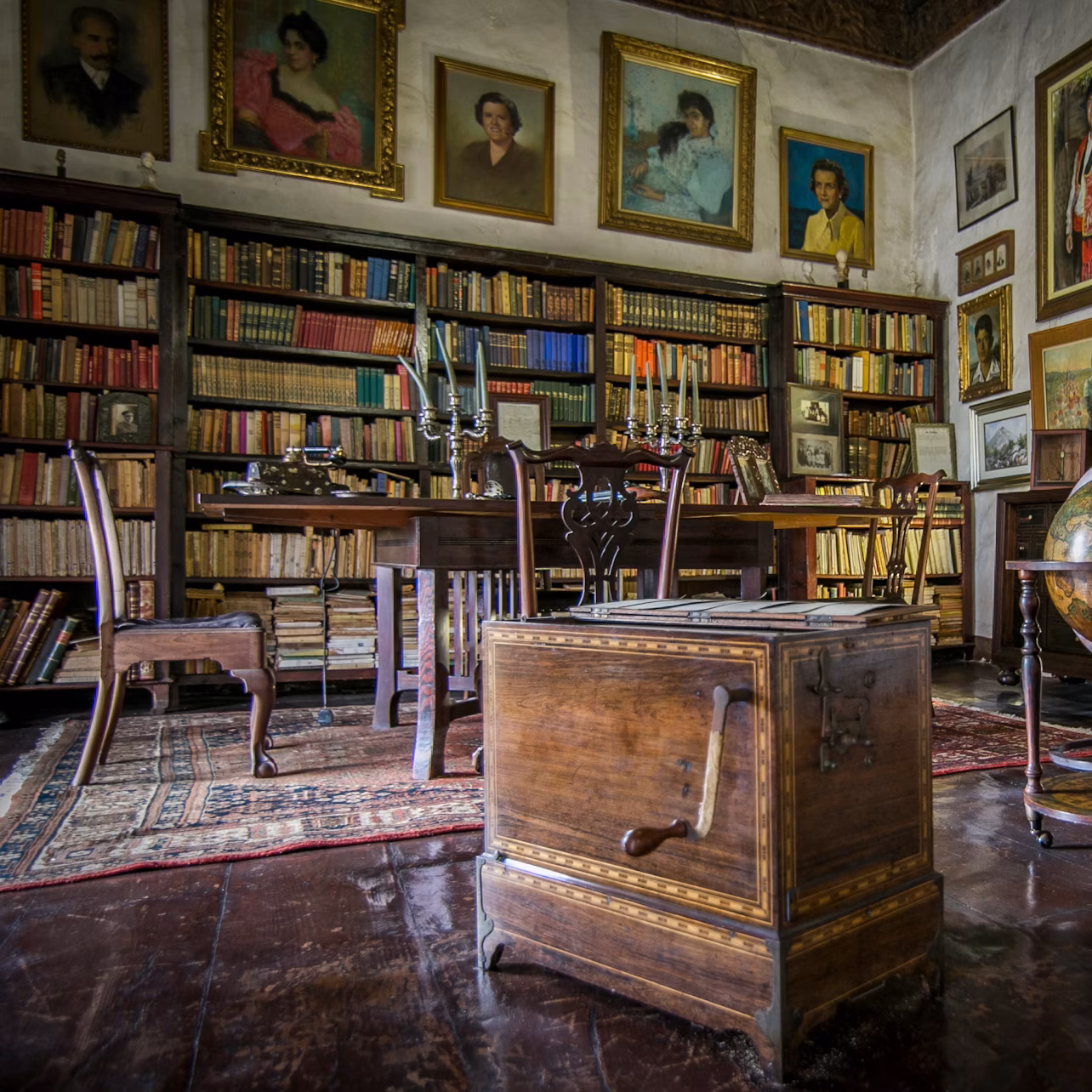 A library with wooden bookshelves, a wooden table with candlesticks, chairs, a globe, and portraits on the wall.
