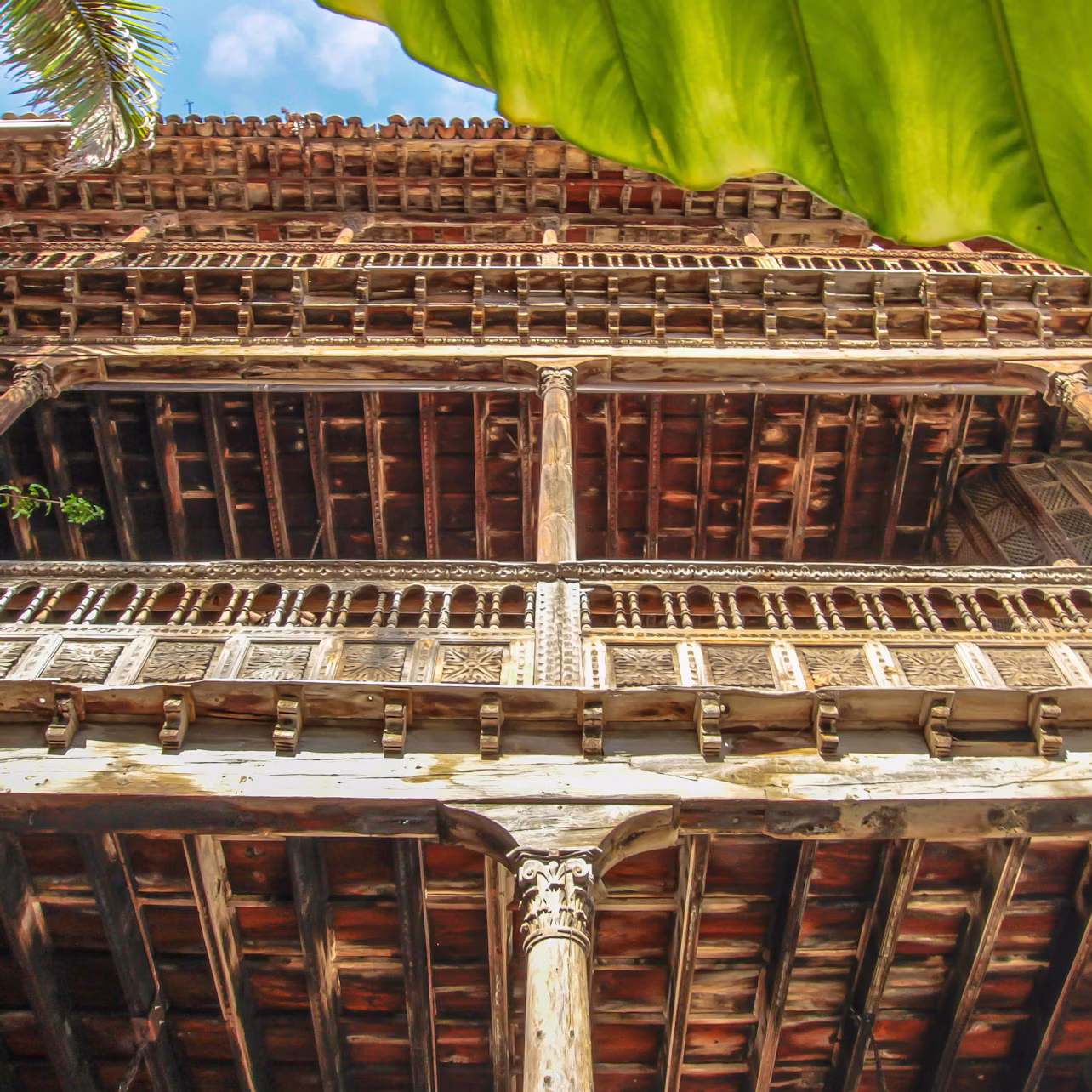 View from below of an intricate, multi-story wooden building with balconies, framed by a large green leaf and a palm frond.