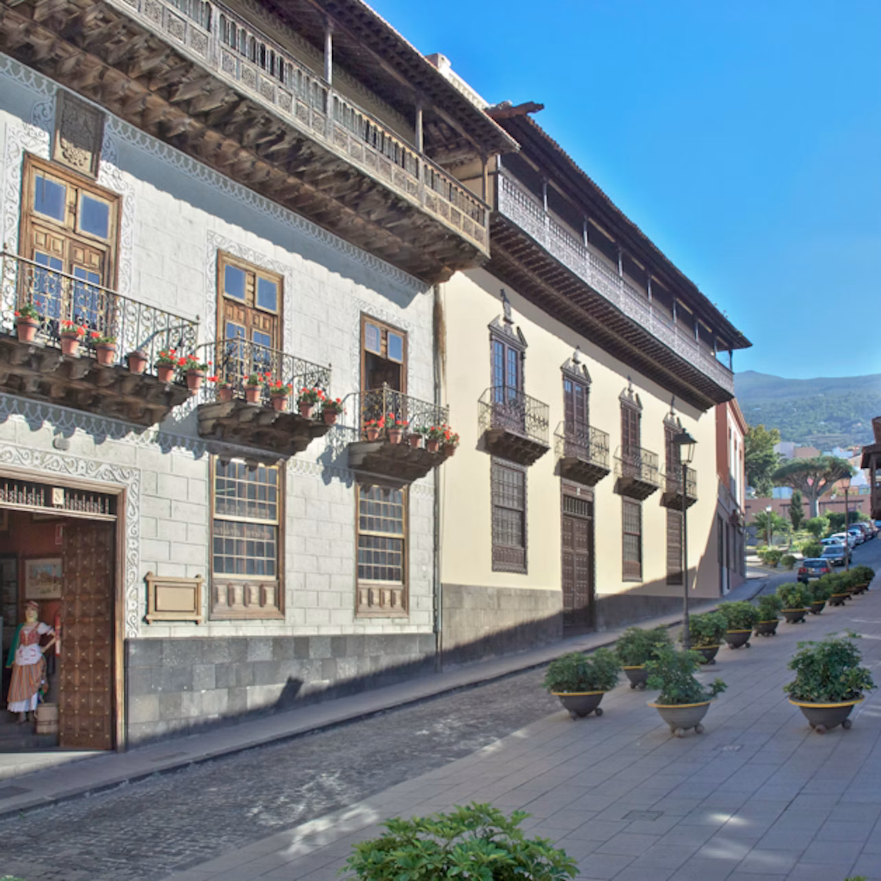 A street with historic buildings featuring wooden balconies and flower pots. Potted plants line the sidewalk under a clear blue sky.