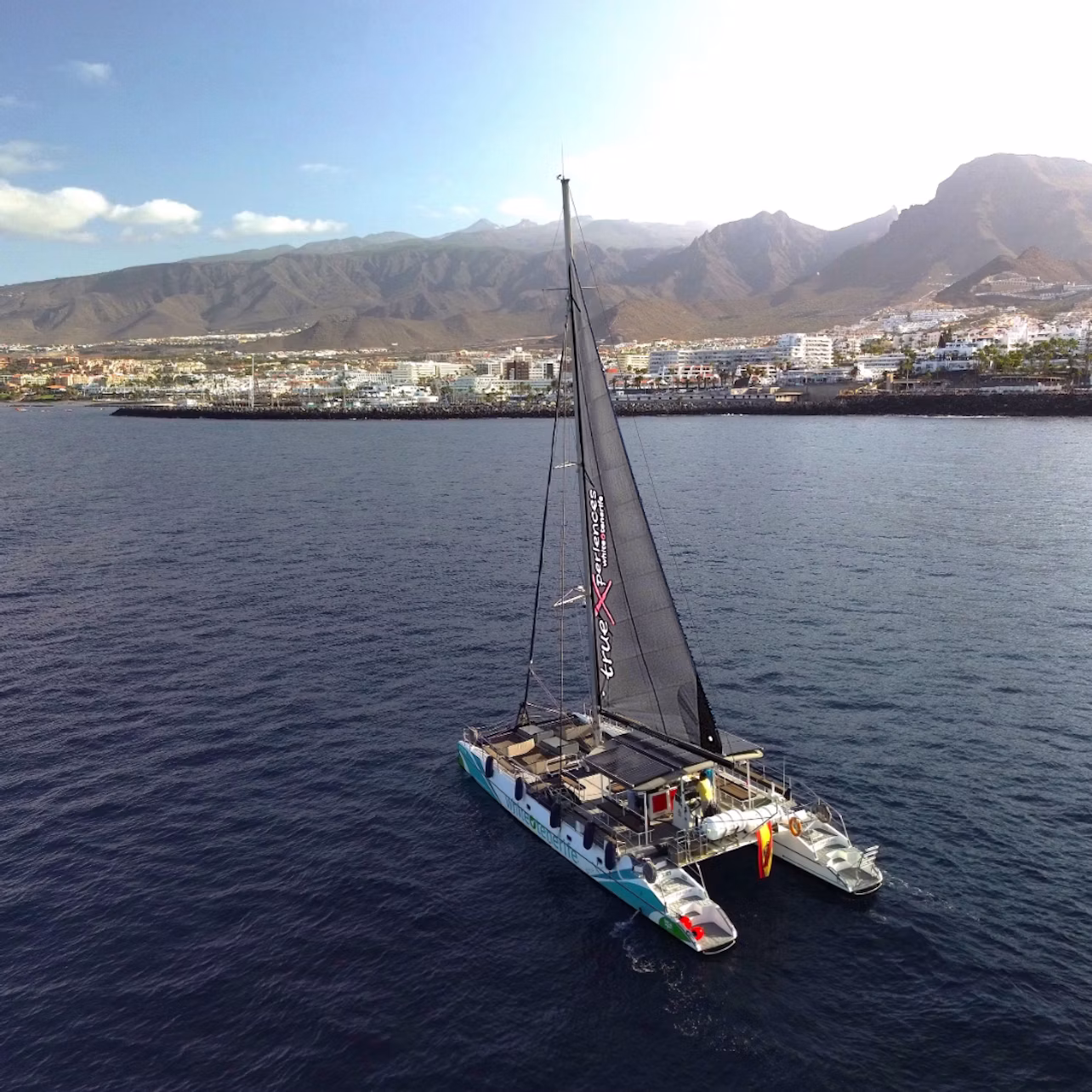 A catamaran with a single black sail on calm waters, with a coastal town and mountain range in the background under a blue sky.