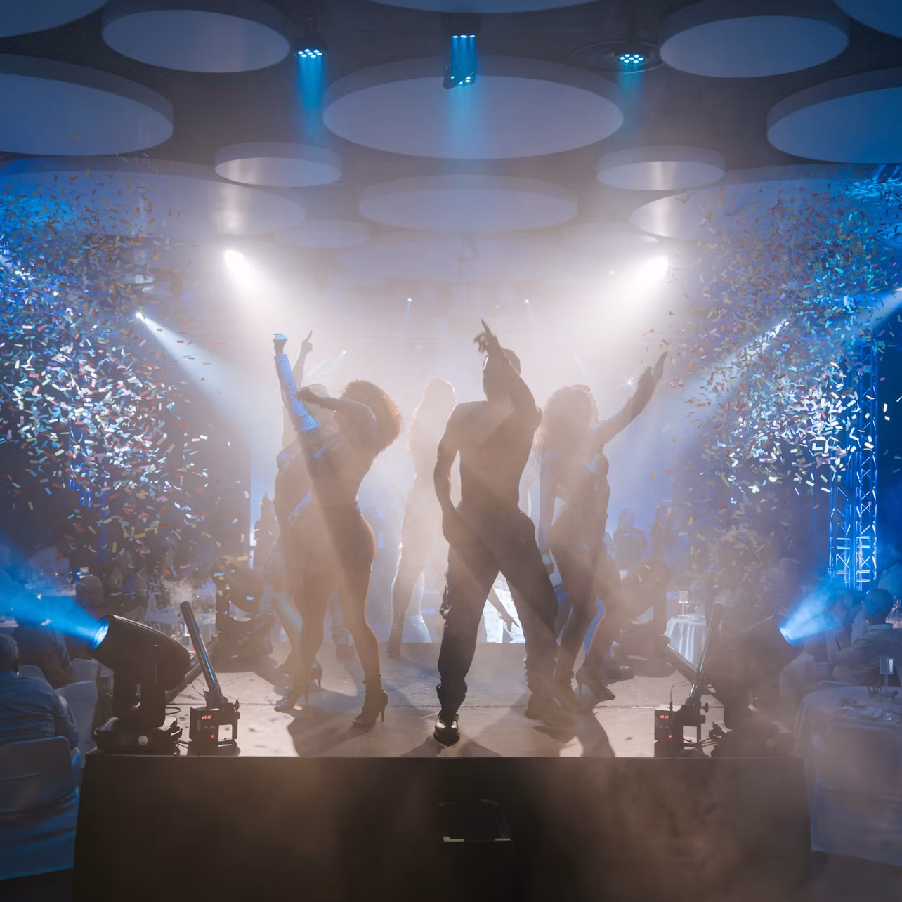 Silhouetted dancers performing on a stage with confetti falling and dramatic stage lighting in a crowded indoor venue.