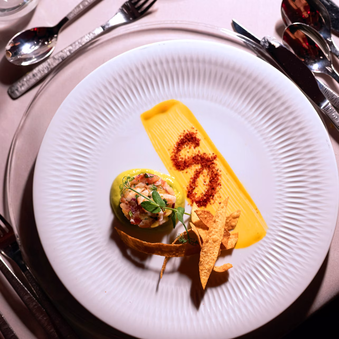 A white plate with a gourmet dish, featuring yellow sauce, decorative herbs, and crispy elements, on a set dining table.
