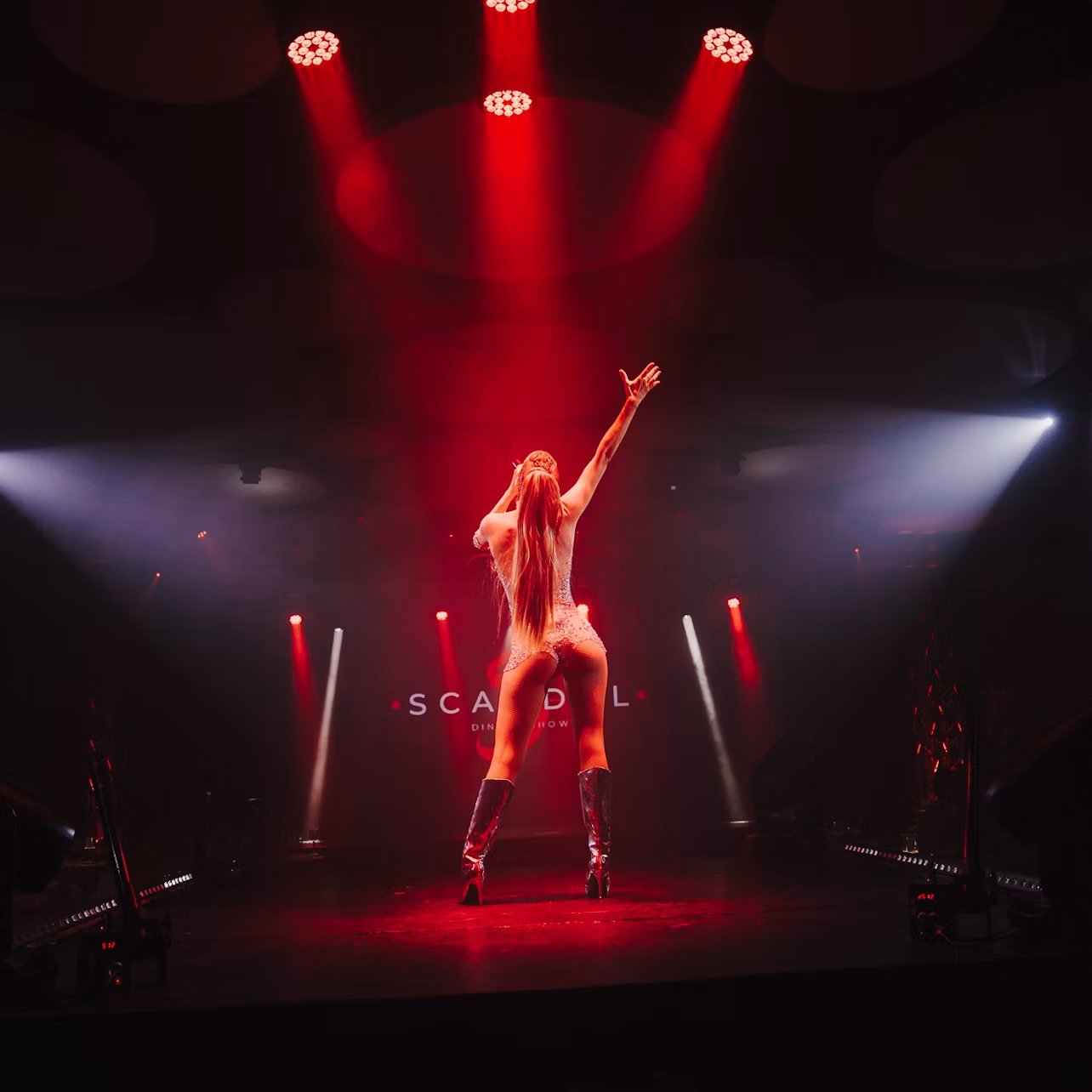 Performer on stage under red and white lights, with arms raised, in a dramatic pose during a show.