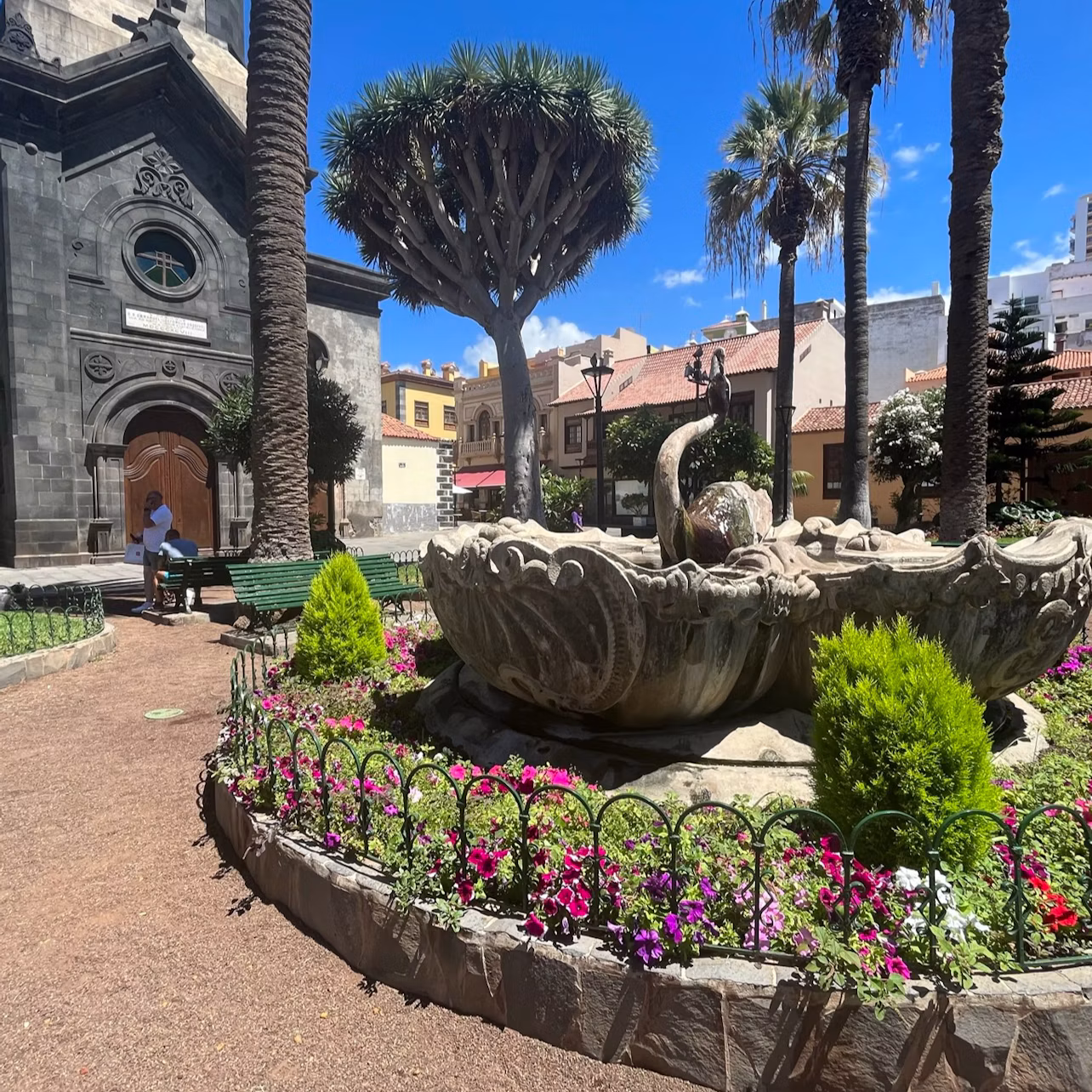 A garden with a stone fountain, flowers, and trees, adjacent to a dark stone building with an arched doorway under a blue sky.