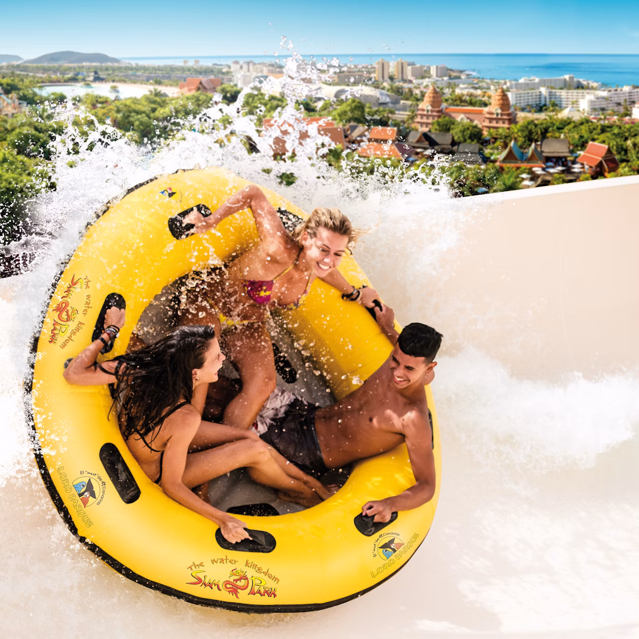 Three people on a yellow inflatable raft ride down a water slide with splashing water, cityscape and ocean in the background.
