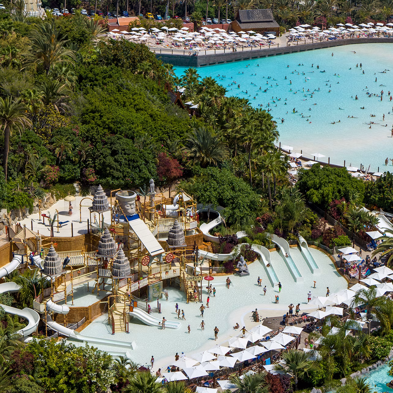 Aerial view of a water park with elaborate slides, pools, and lounging areas; a large pool with numerous swimmers in the background.