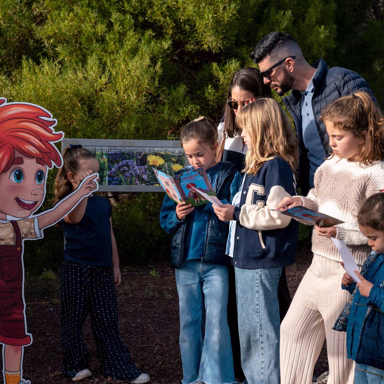 A group of children and an adult stand outdoors reading books near a colorful sign. Trees are visible in the background.