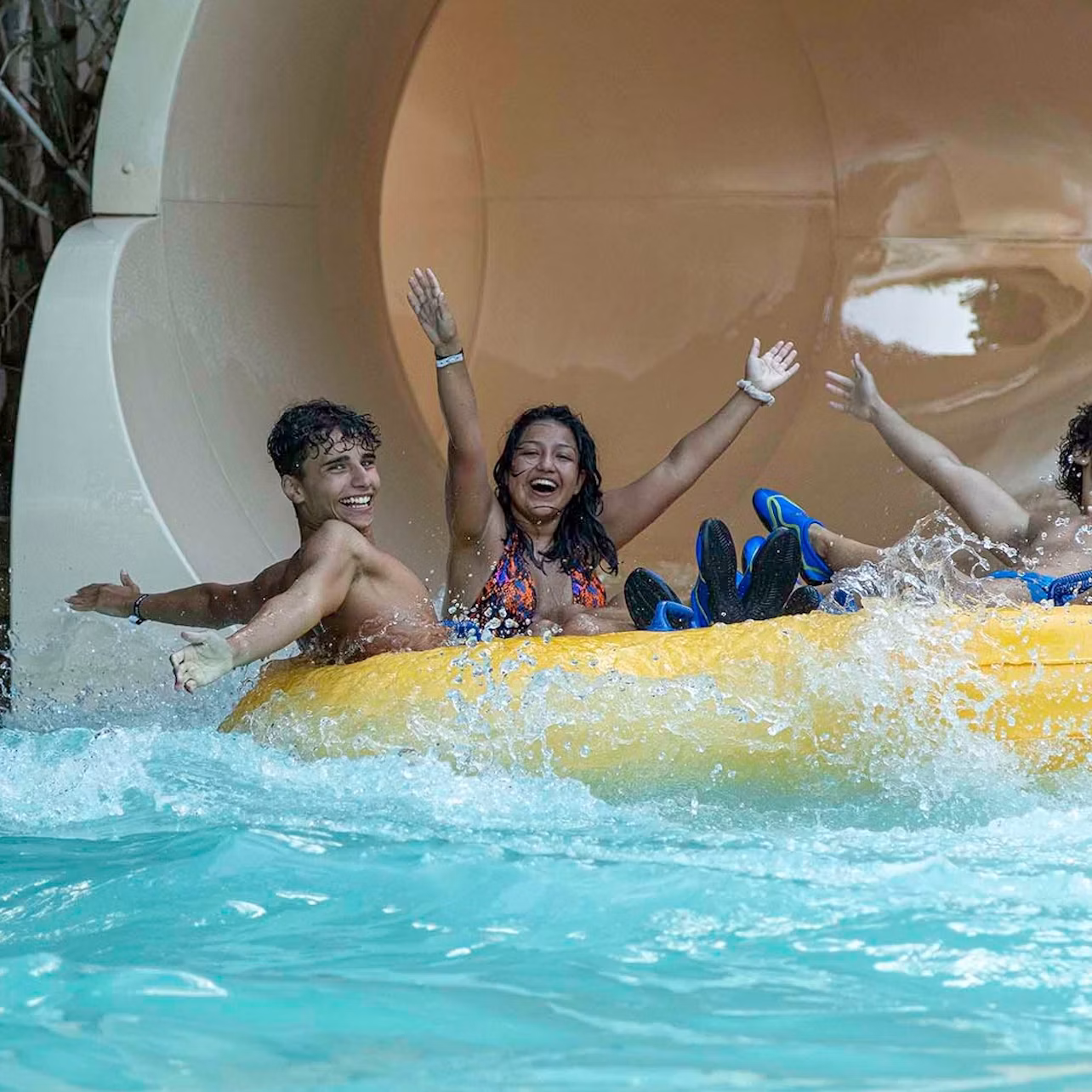 Three people joyfully ride a yellow inflatable raft down a water slide, splashing into the pool below.
