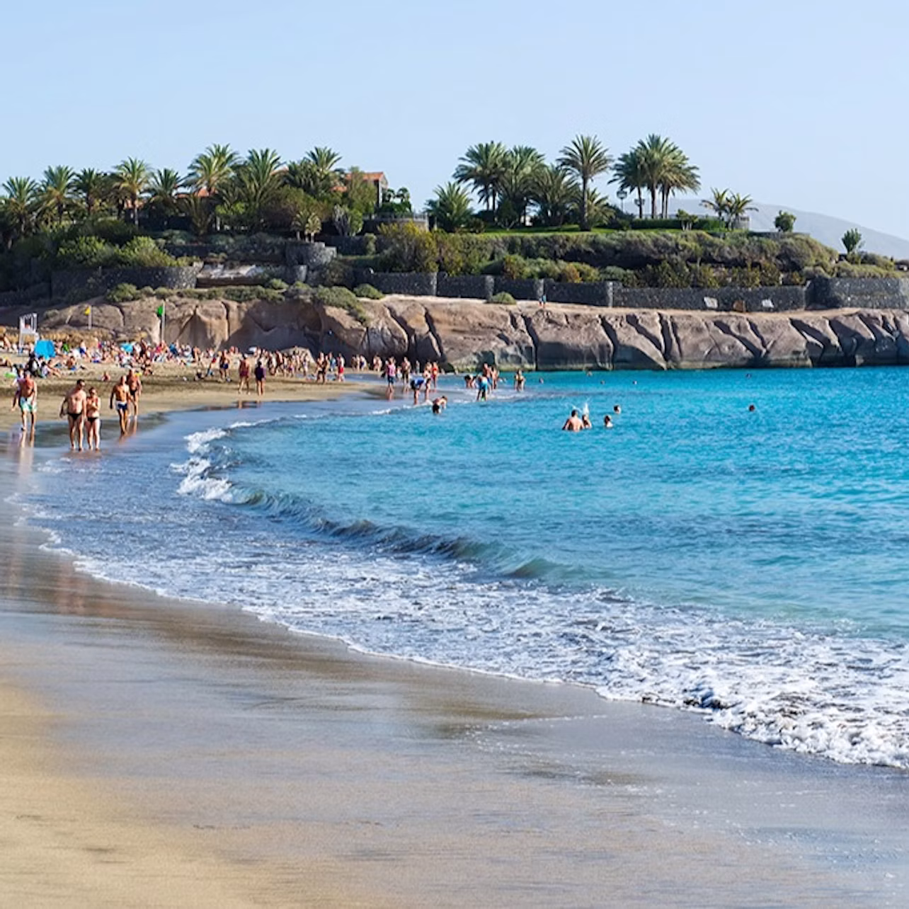 People walking and swimming at a sandy beach with clear blue water, rocky coastline, and palm trees in the background.