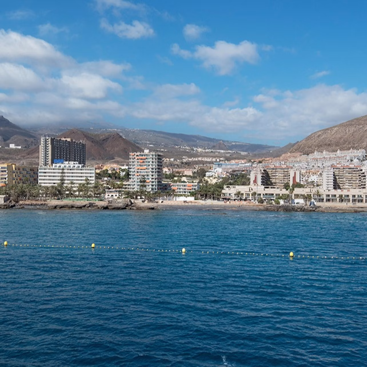 City skyline with buildings along a coastal area, blue sea in the foreground, and mountains in the background under a partly cloudy sky.