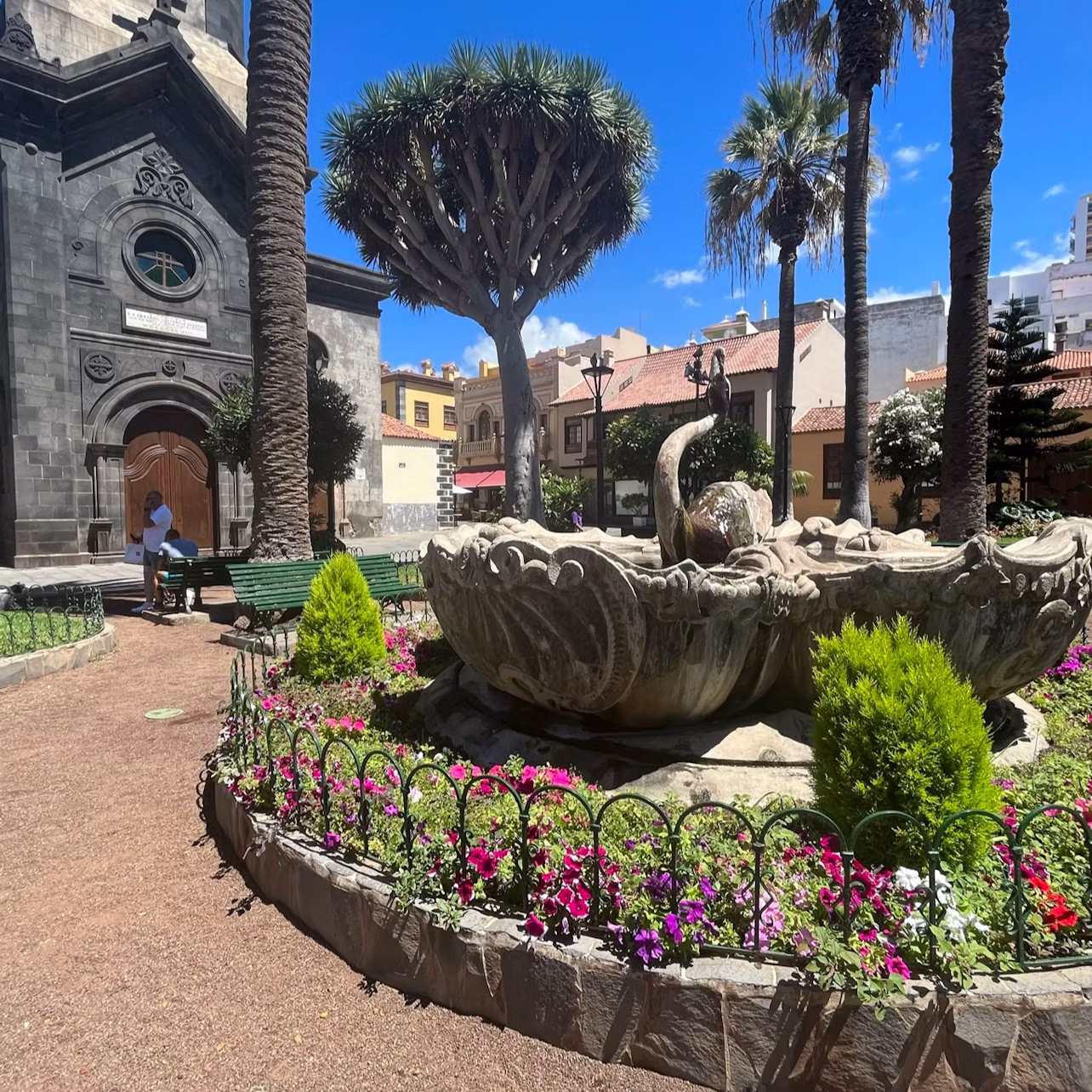 A garden with a stone fountain, flowers, and trees, adjacent to a dark stone building with an arched doorway under a blue sky.