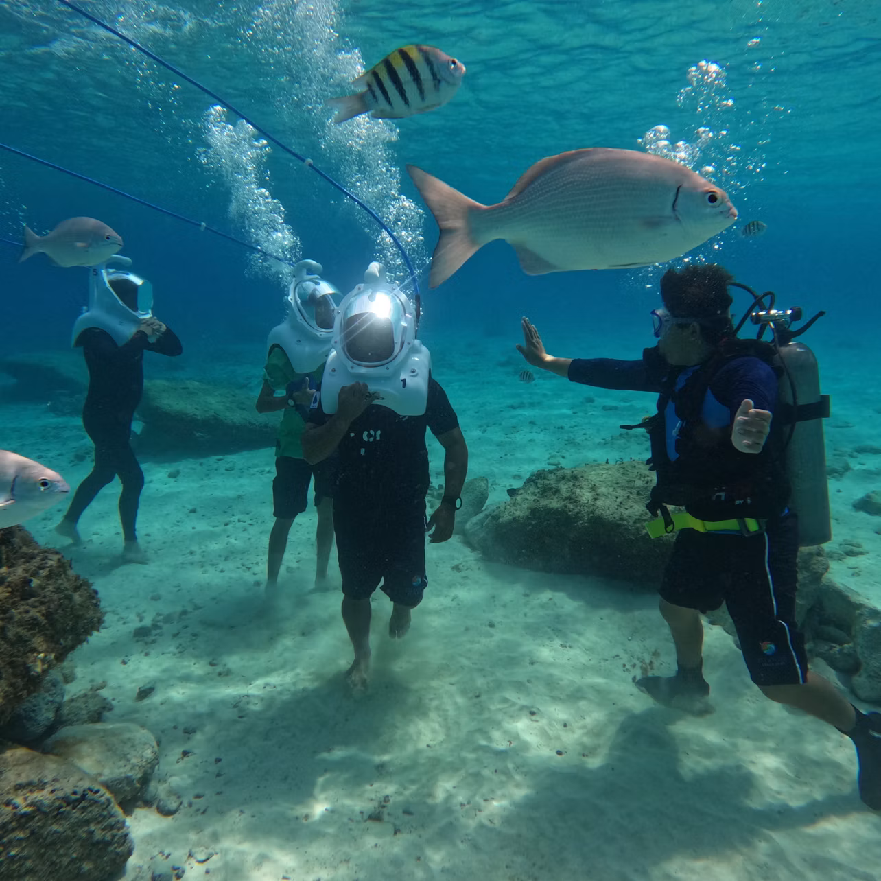 Group of people underwater wearing helmets, surrounded by fish and a scuba diver giving instructions. Clear blue water, rocky seabed.