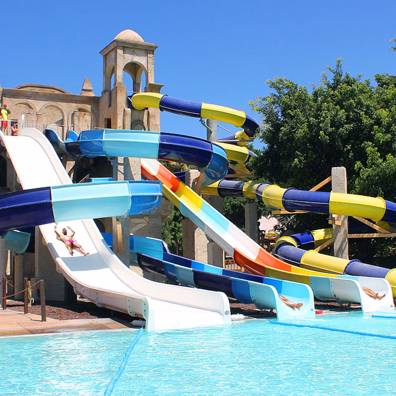 Multiple colorful water slides leading into a large pool with people sliding down, surrounded by trees and a building in the background.