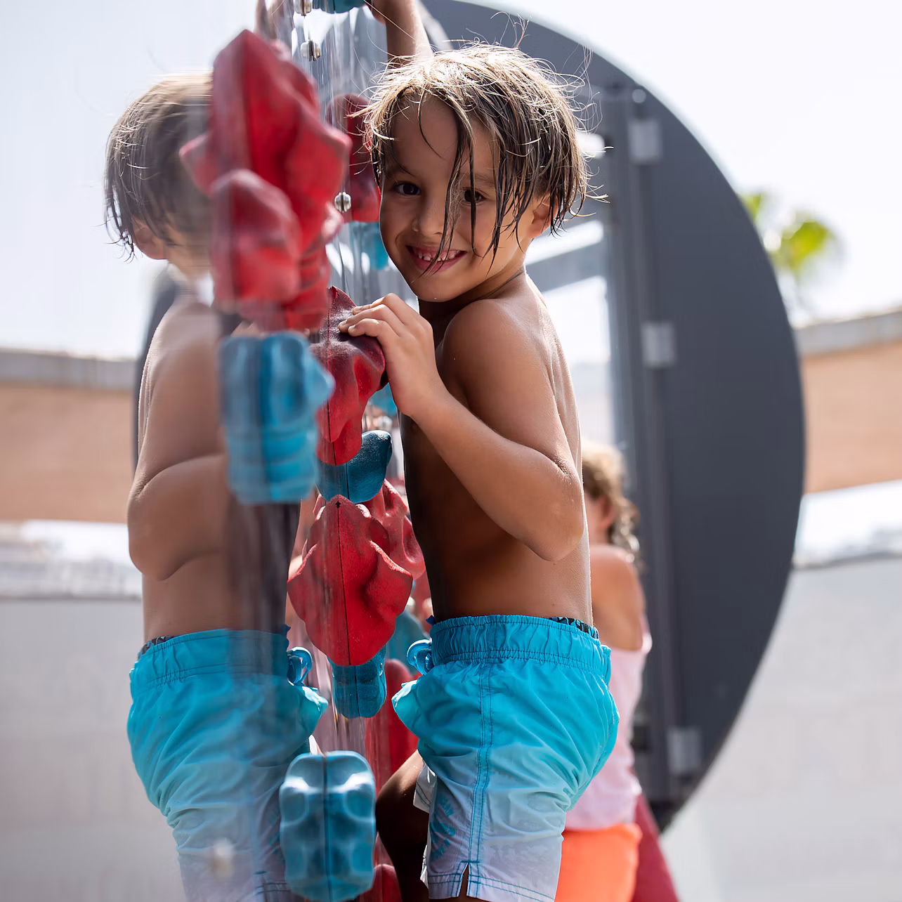 A child in blue shorts climbs on a playground structure, smiling towards the camera, with another child in the background.