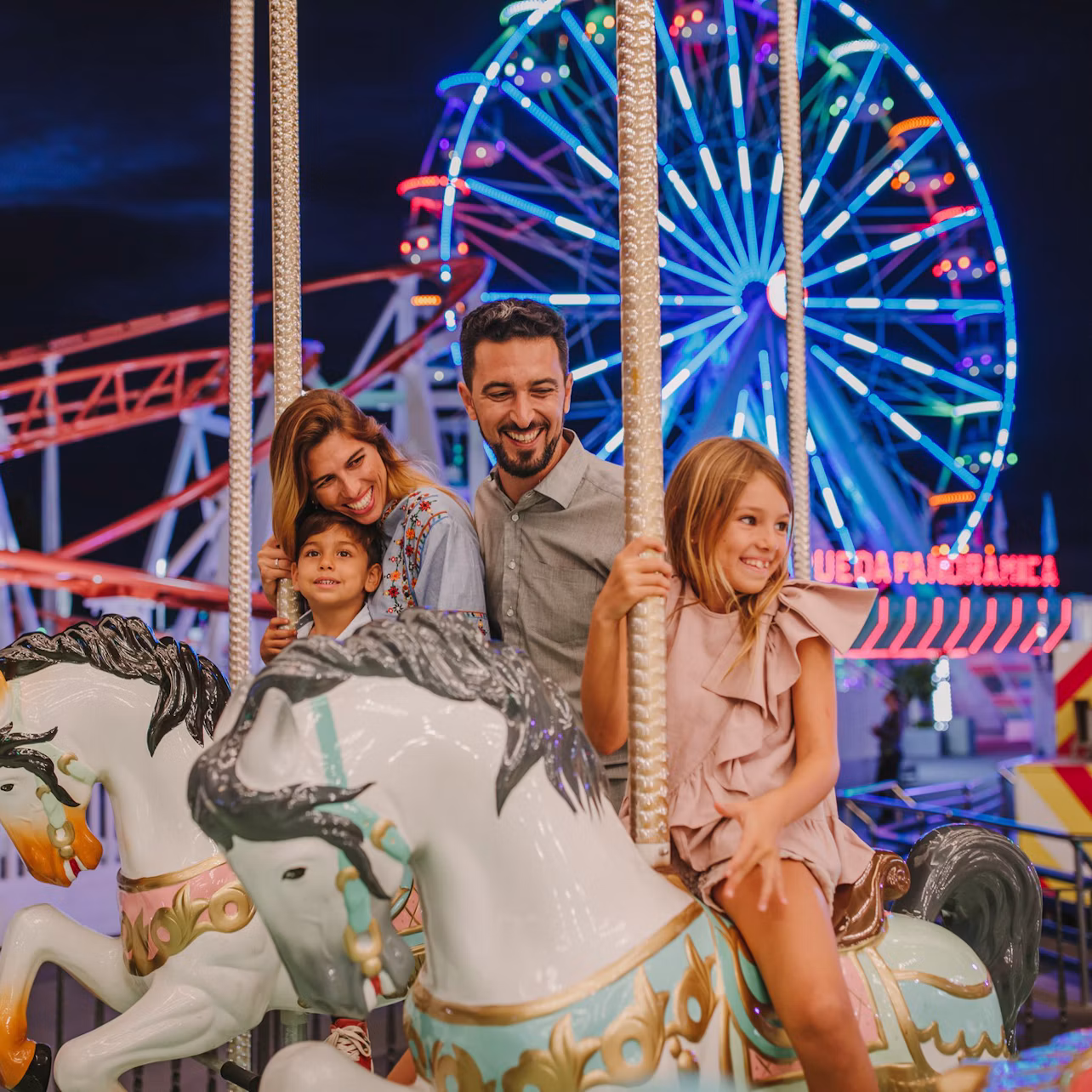 A family enjoying a carousel ride at night with a lit-up Ferris wheel and rollercoaster in the background.
